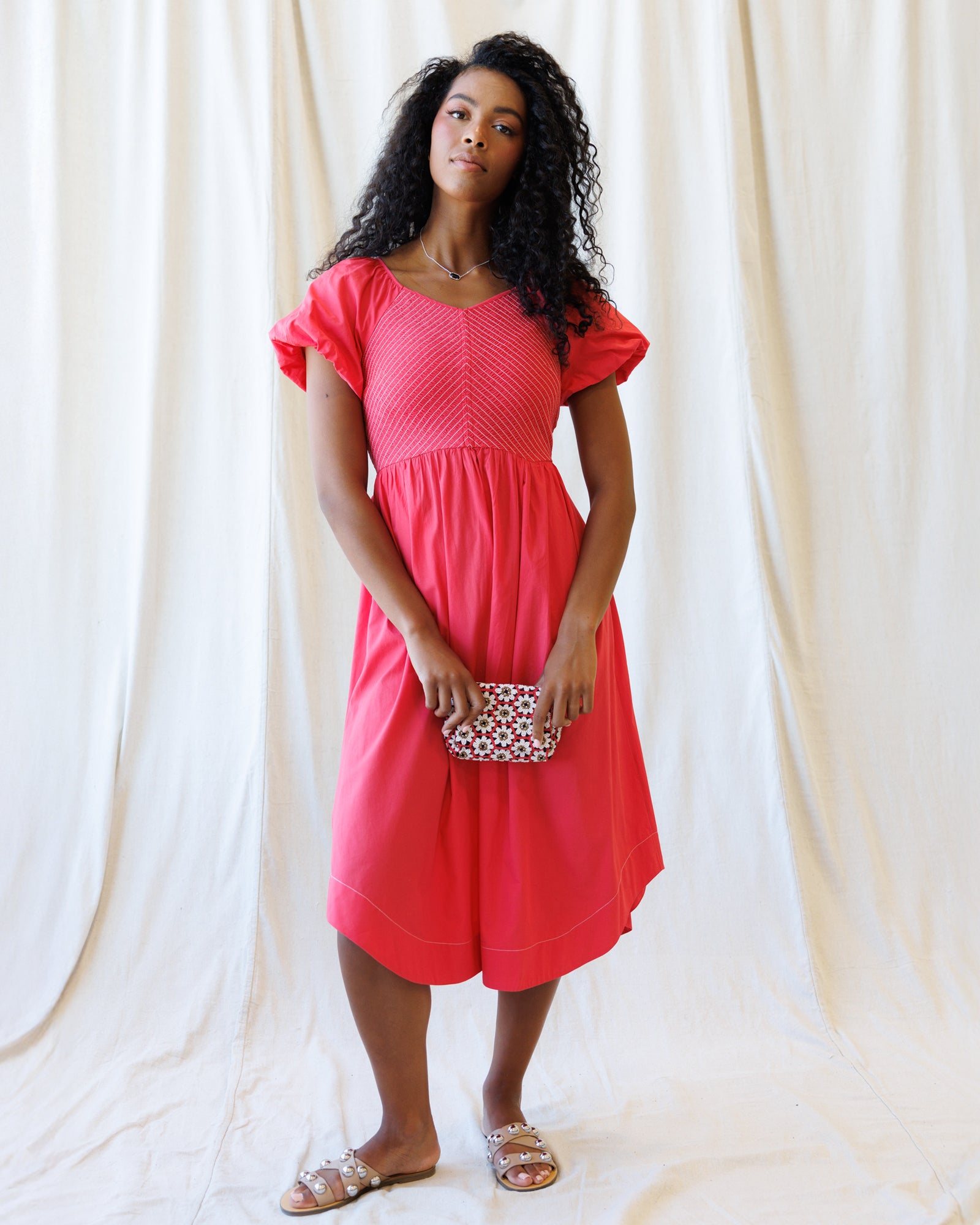 Woman wearing a pink dress holding a clutch against a white curtain background