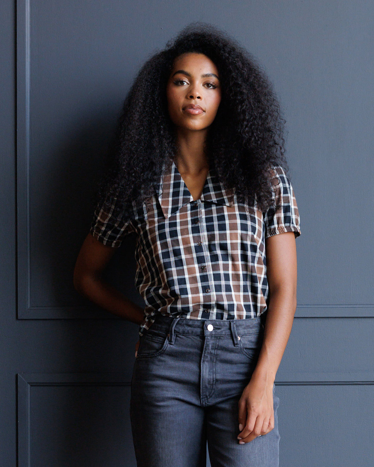 A woman with voluminous dark curly hair stands against a dark blue paneled wall, wearing a short-sleeve button-up plaid shirt in navy, white, and brown, paired with dark gray jeans.
