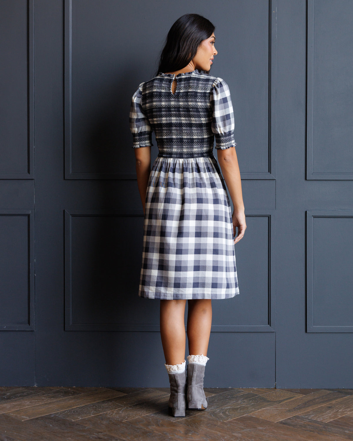 A woman stands with her back to the camera wearing a knee-length plaid dress with puff sleeves and a back keyhole detail, paired with gray socks and boots, in front of a dark paneled wall.