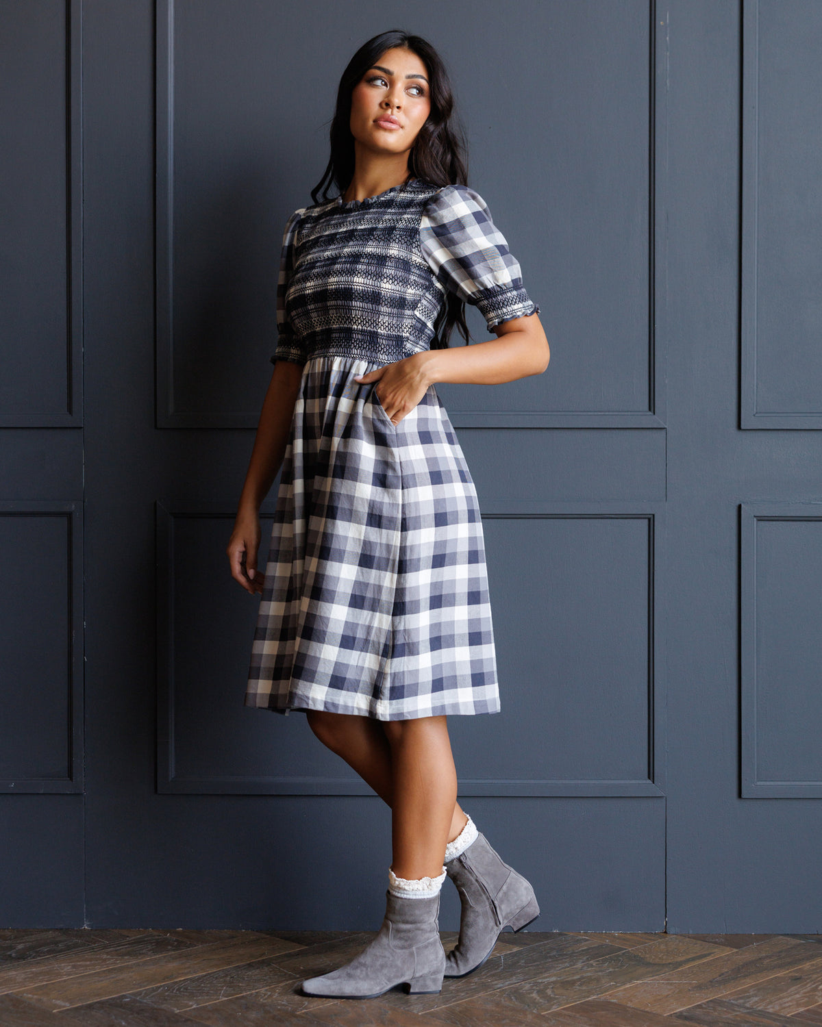 A woman models a short-sleeved plaid dress with a navy, white, and gray checkered pattern, paired with gray suede ankle boots, standing against a dark blue paneled wall.