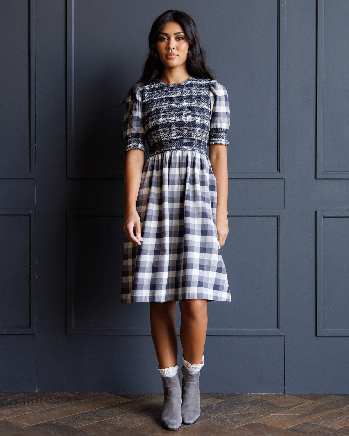 A woman stands against a dark blue paneled wall wearing a knee-length plaid dress with puff sleeves, a smocked waist, and gray ankle boots.