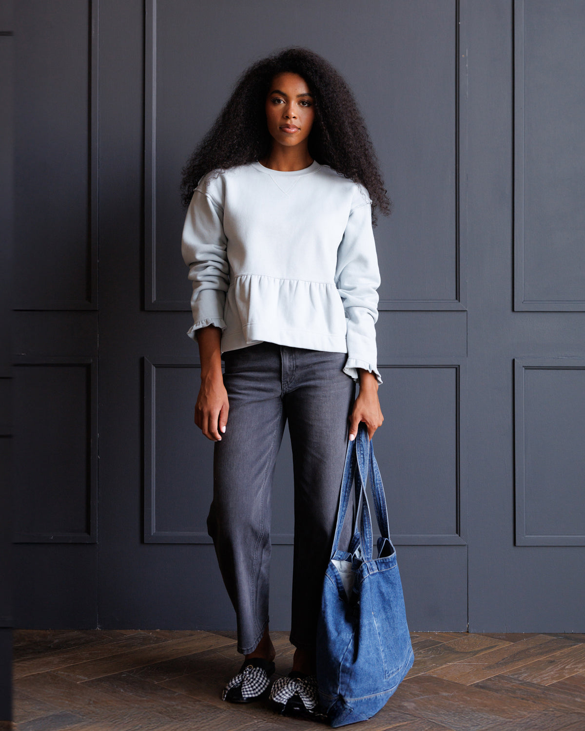 A woman stands in front of a dark paneled wall wearing a light blue long-sleeve peplum top, dark gray pants, and holding a large blue denim tote bag.