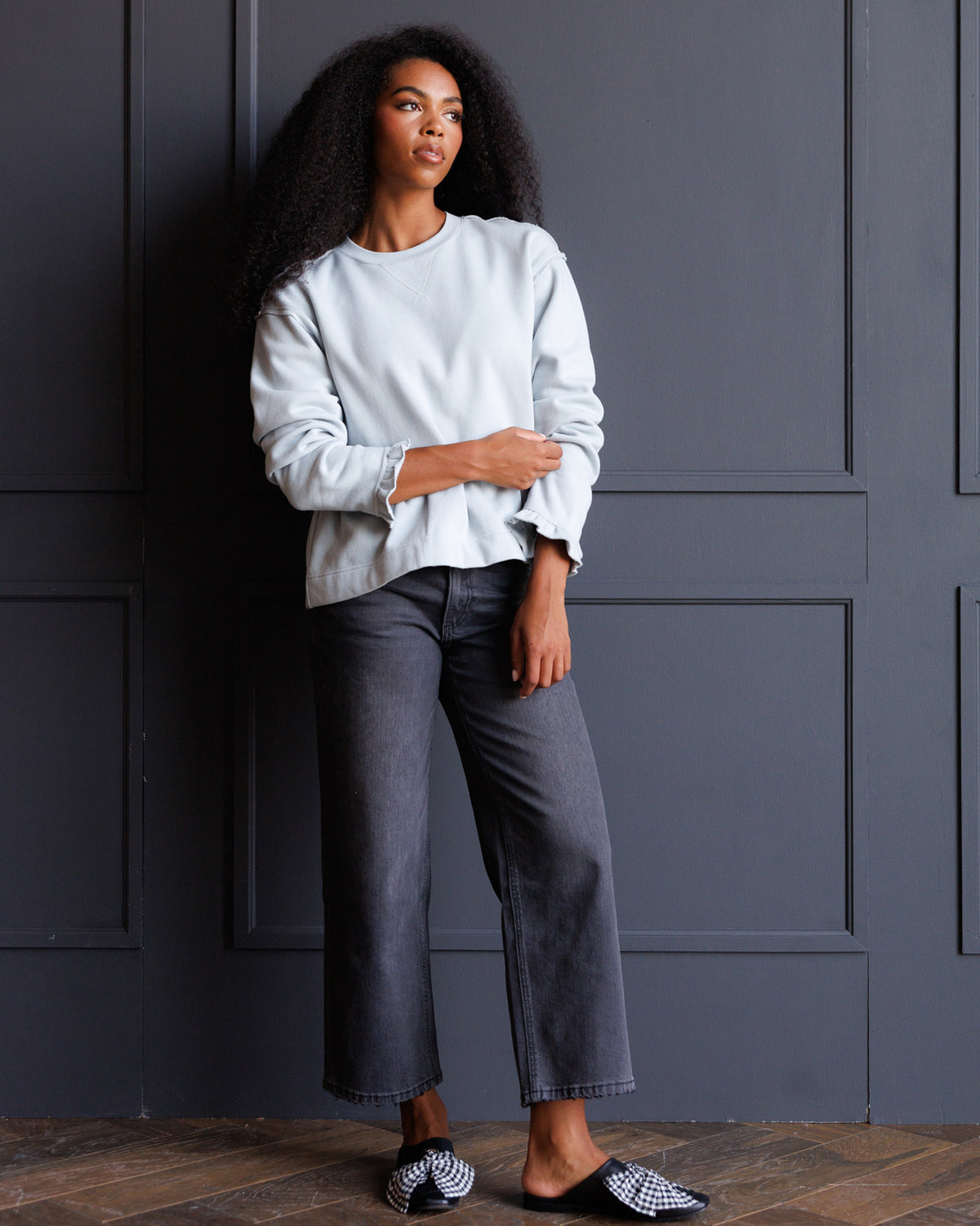 A woman stands against a dark gray paneled wall wearing a light blue long-sleeve top, dark gray cropped jeans, and black-and-white patterned slip-on shoes.