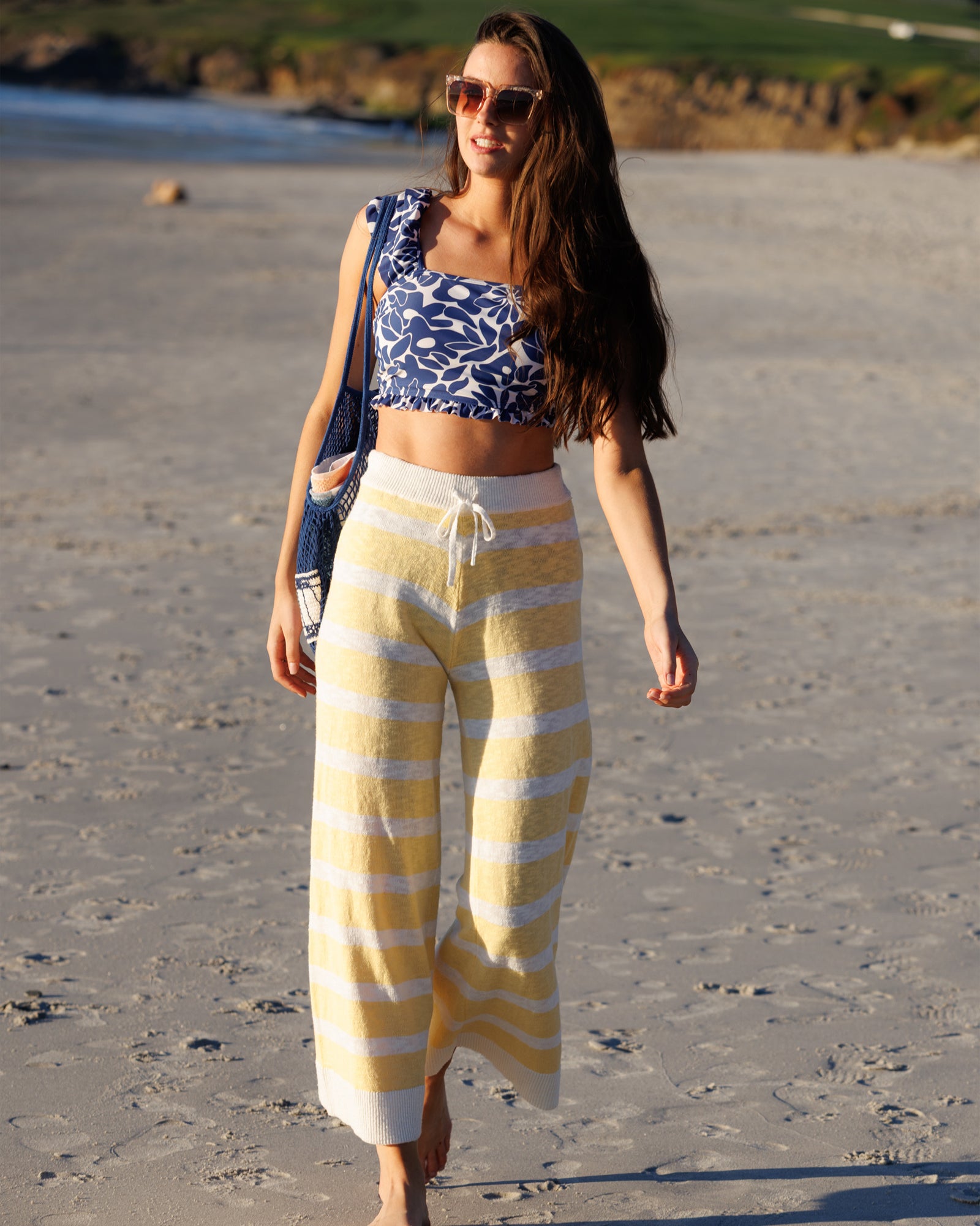 Woman in a blue and white patterned top and yellow and white striped pants walking on a sandy beach.