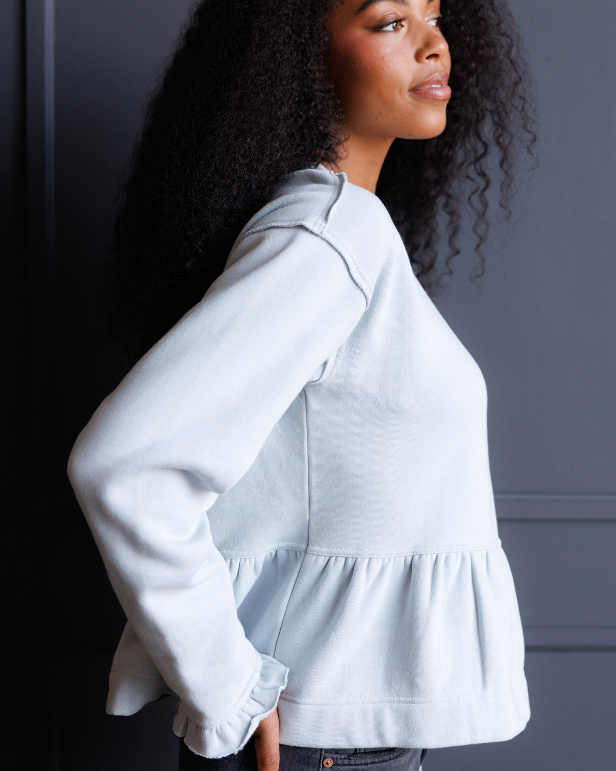 A woman wearing a light blue long-sleeve cropped sweatshirt with a ruffled hem, shown in a side profile against a dark gray paneled wall.