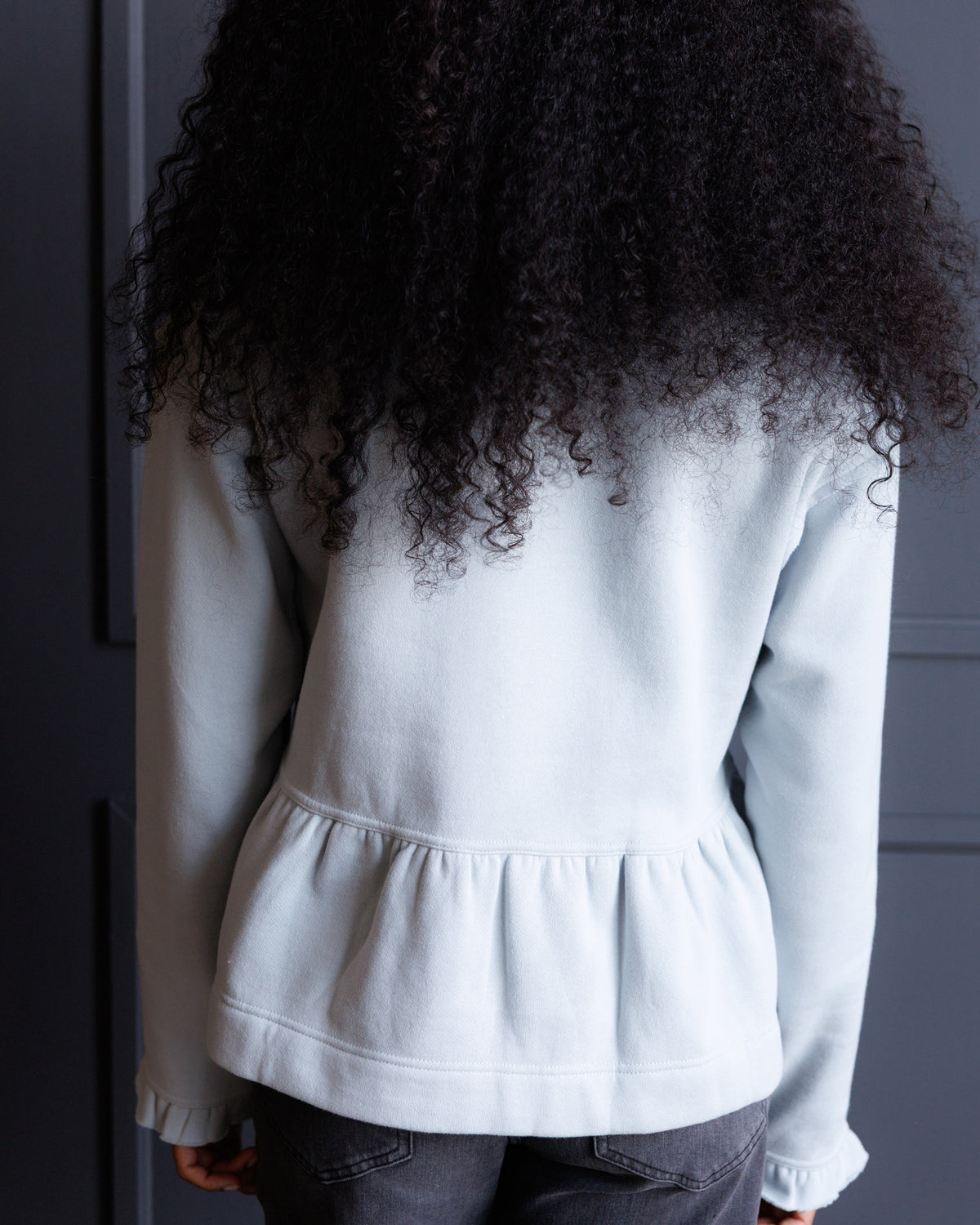 A person shown from the back wearing a white long-sleeve top with a gathered peplum waist seam, paired with dark gray jeans, against a dark gray paneled door.