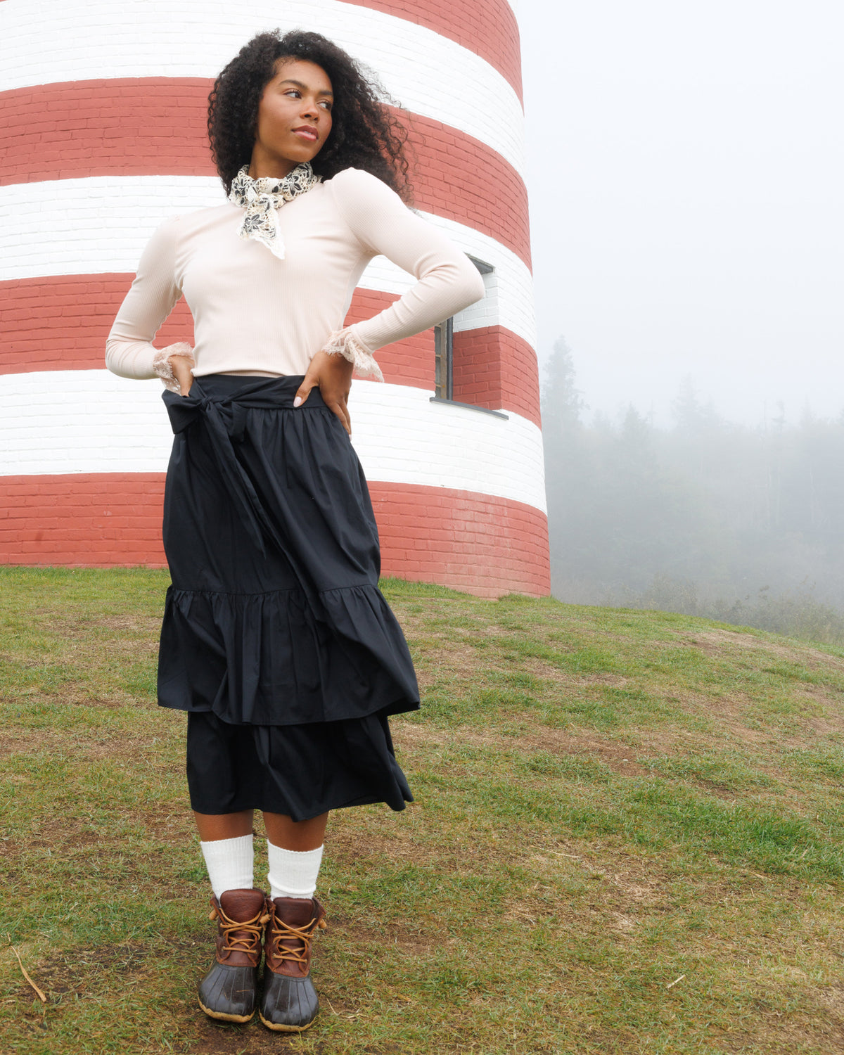 A woman stands outdoors in front of a red-and-white striped lighthouse on a grassy hill, wearing a cream long-sleeve top, a black tiered skirt, white socks, and brown lace-up boots.