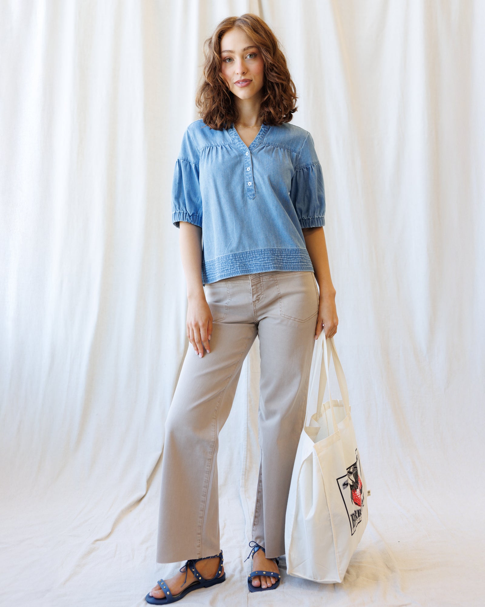 Woman wearing a blue top and light gray pants, holding a white tote bag with a design, against a white curtain background.