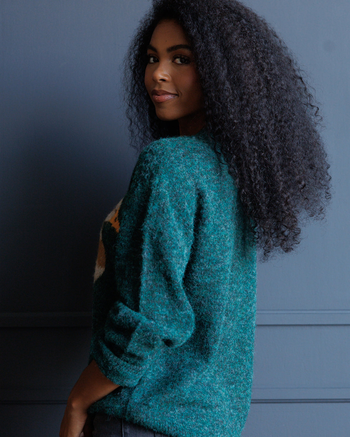 A woman with long, dark, curly hair is shown from a side-back angle wearing a textured teal-green knit sweater against a dark blue-gray paneled wall.
