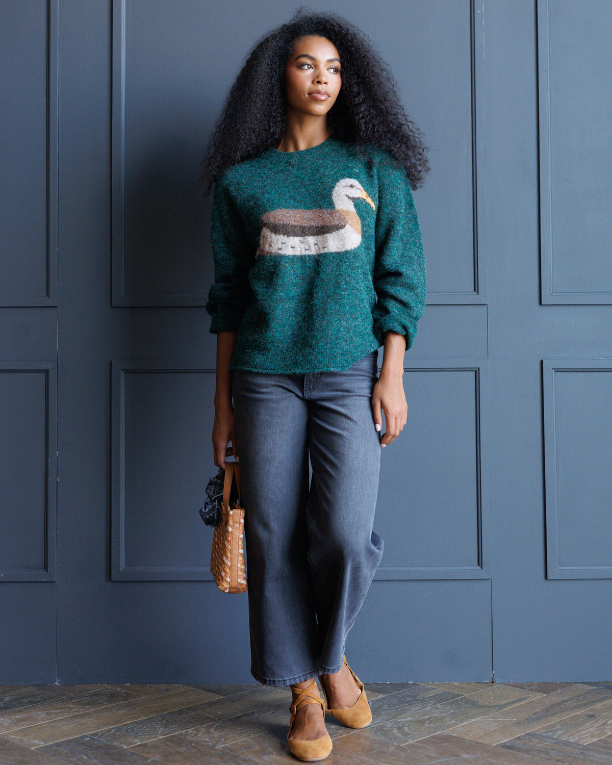 A woman stands against a dark blue paneled wall wearing a teal knit sweater with a large duck motif on the chest, blue jeans, tan loafers, and holding a woven tan handbag.