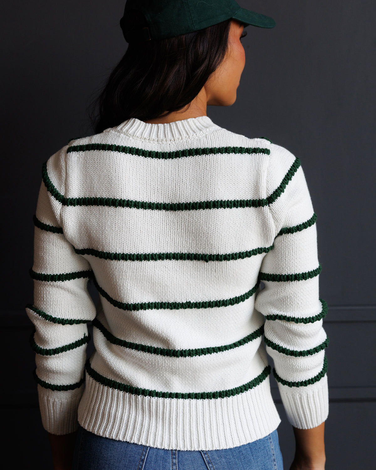 A person shown from the back wearing a white knit sweater with dark green horizontal stripes and a matching dark green cap, against a dark gray wall.