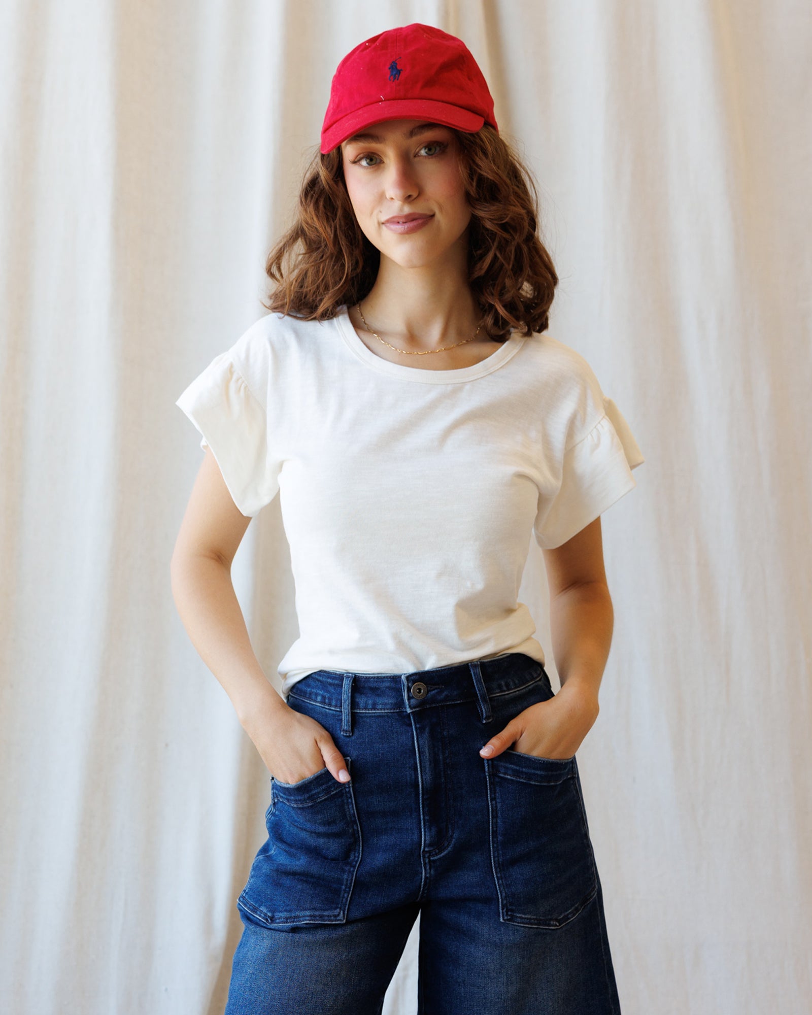 Woman wearing a white t-shirt, blue jeans, and a red cap against a plain background
