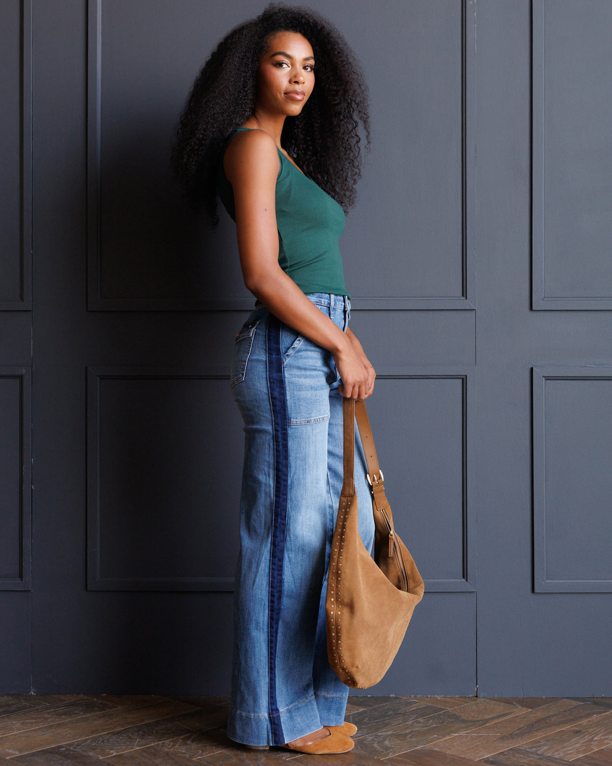 A woman stands in profile against a dark gray paneled wall, wearing a green tank top, wide-leg blue jeans, and tan shoes, while holding a tan suede shoulder bag.