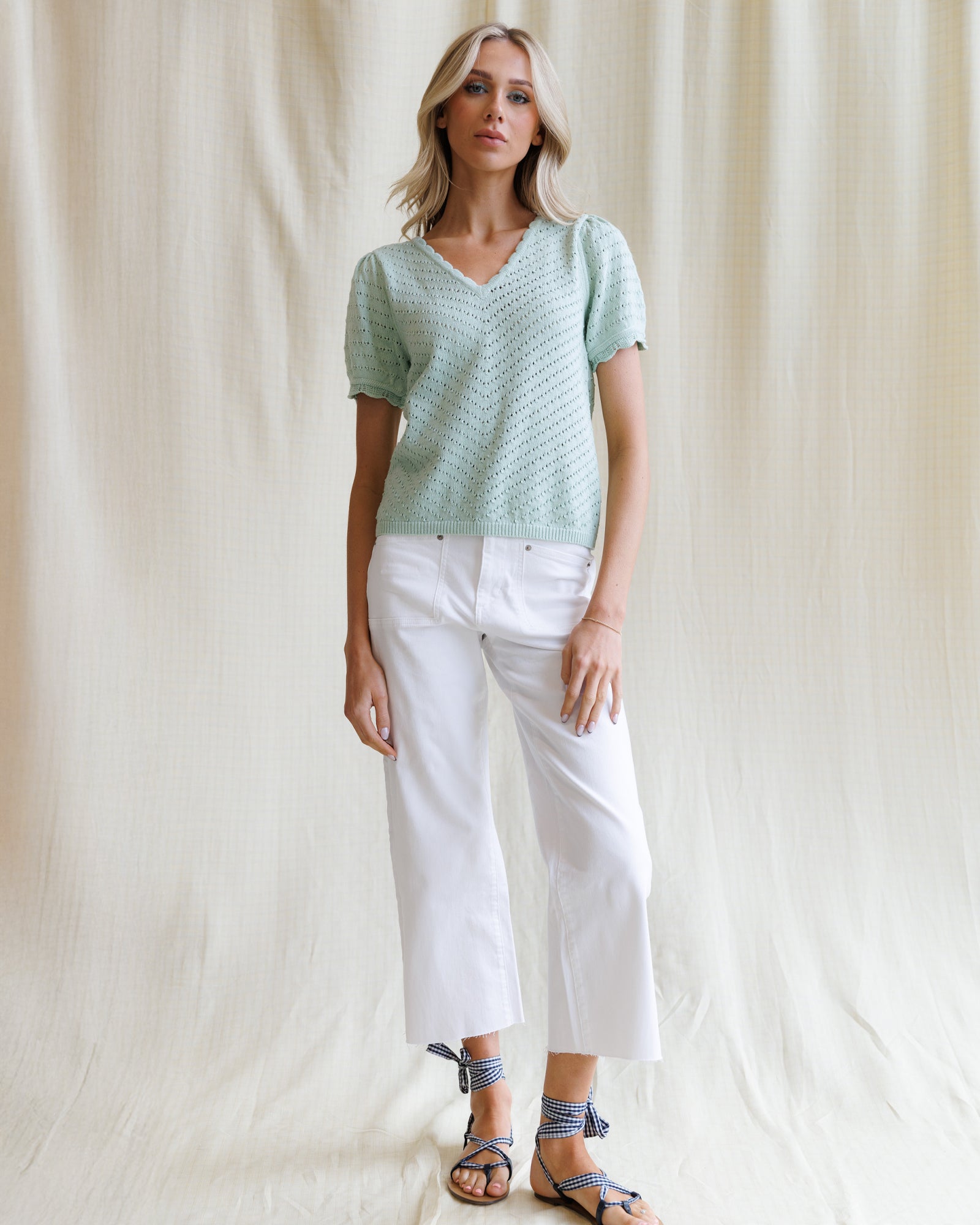 Woman wearing a light green knitted top and white pants against a plain background