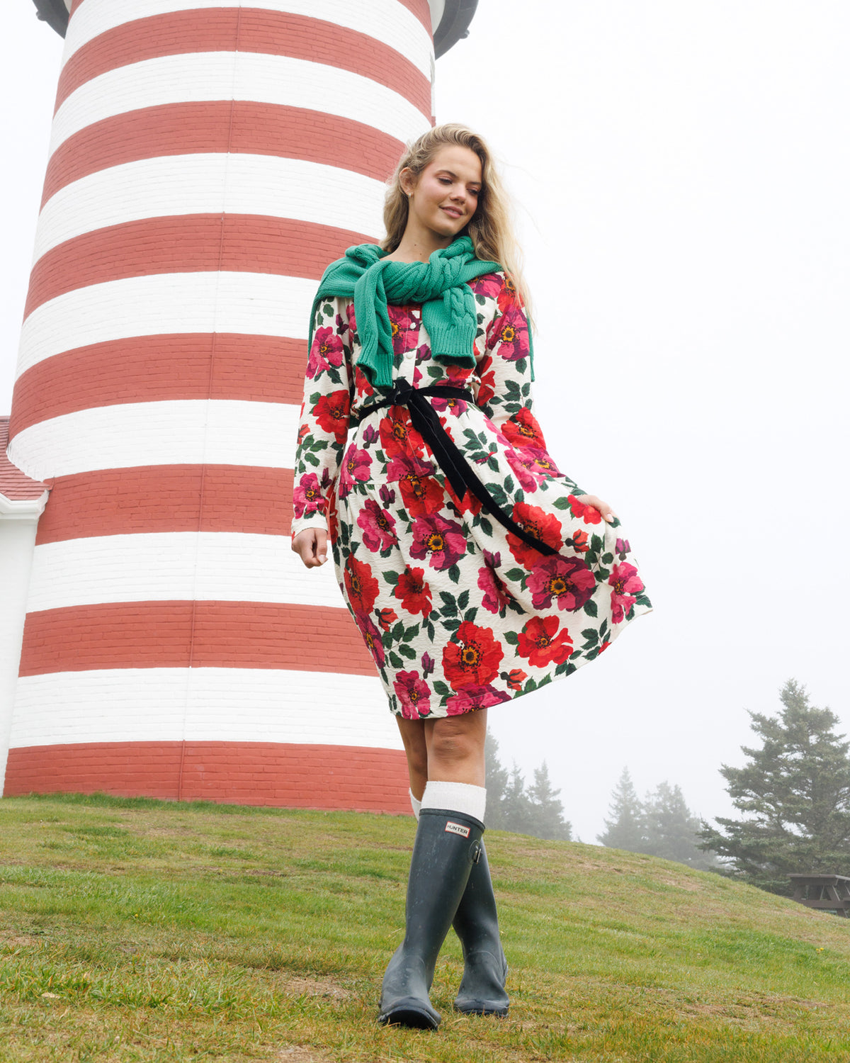 A woman stands outdoors in front of a red-and-white striped lighthouse wearing a white dress with a bold red and pink floral print, a bright green scarf, a black belt, white socks, and dark rain boots.