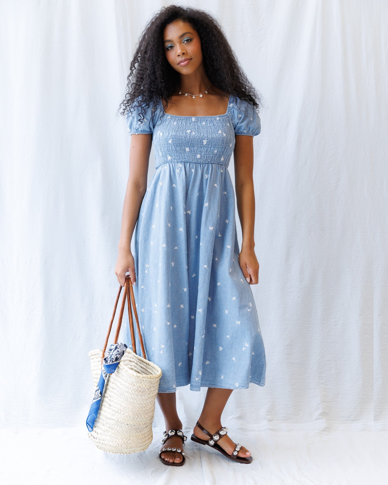 Woman wearing a blue dress with white polka dots, holding a straw bag against a white background