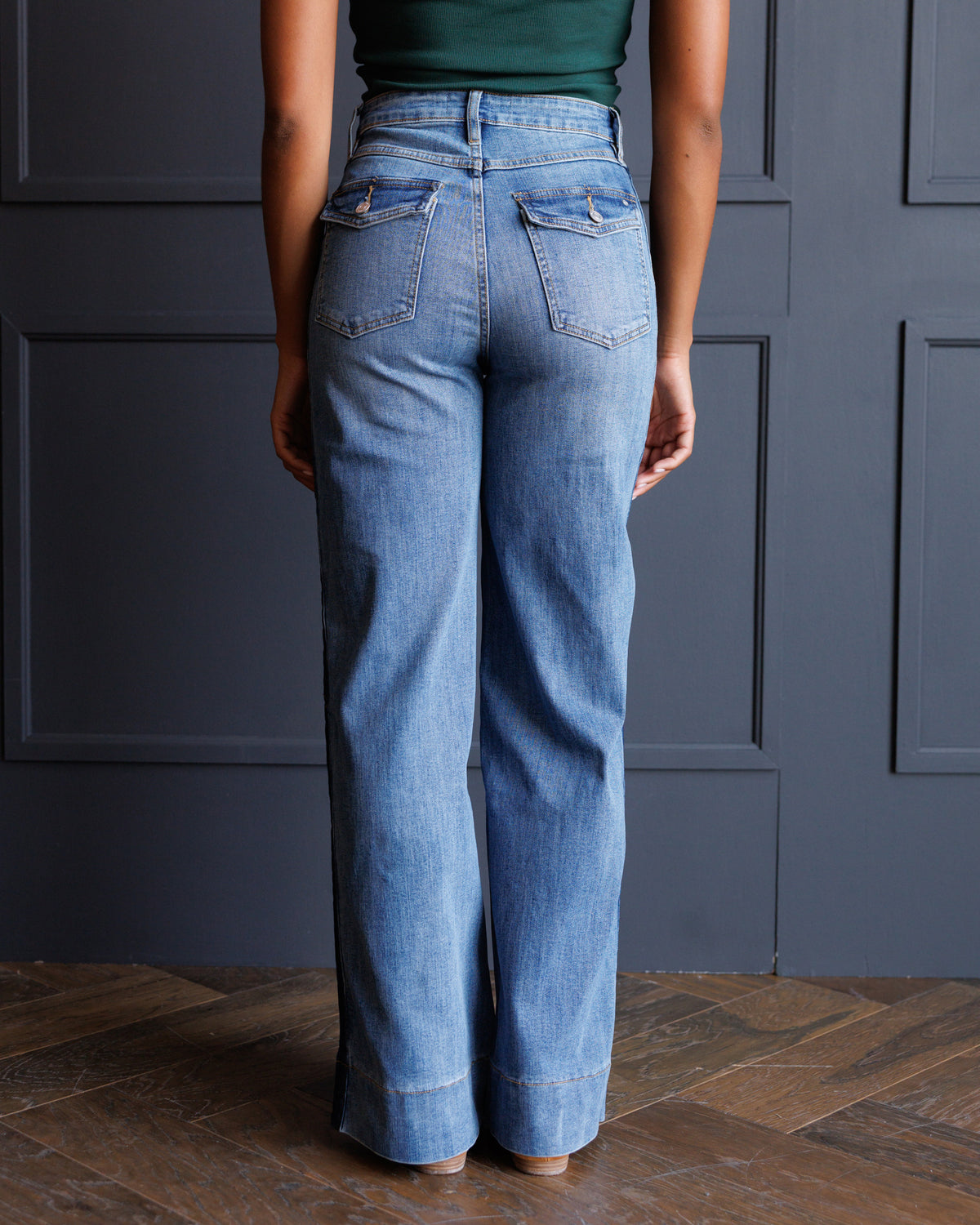 A person shown from the back wearing light blue wide-leg jeans, standing on a wood floor in front of a dark paneled wall.