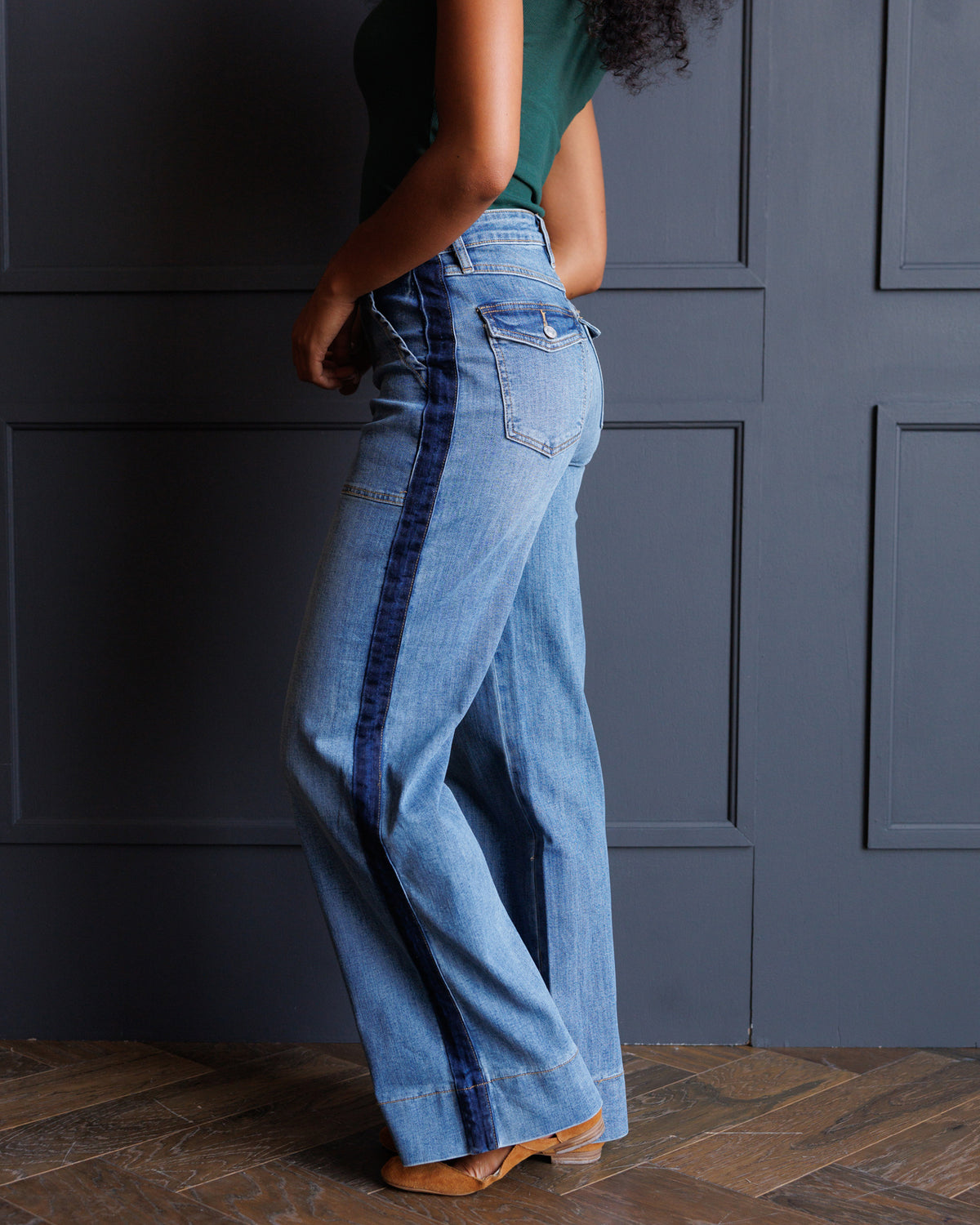 A person wearing light blue wide-leg denim jeans with dark blue vertical stripe detailing down the sides, paired with a dark green top and tan shoes, standing against a dark paneled wall.