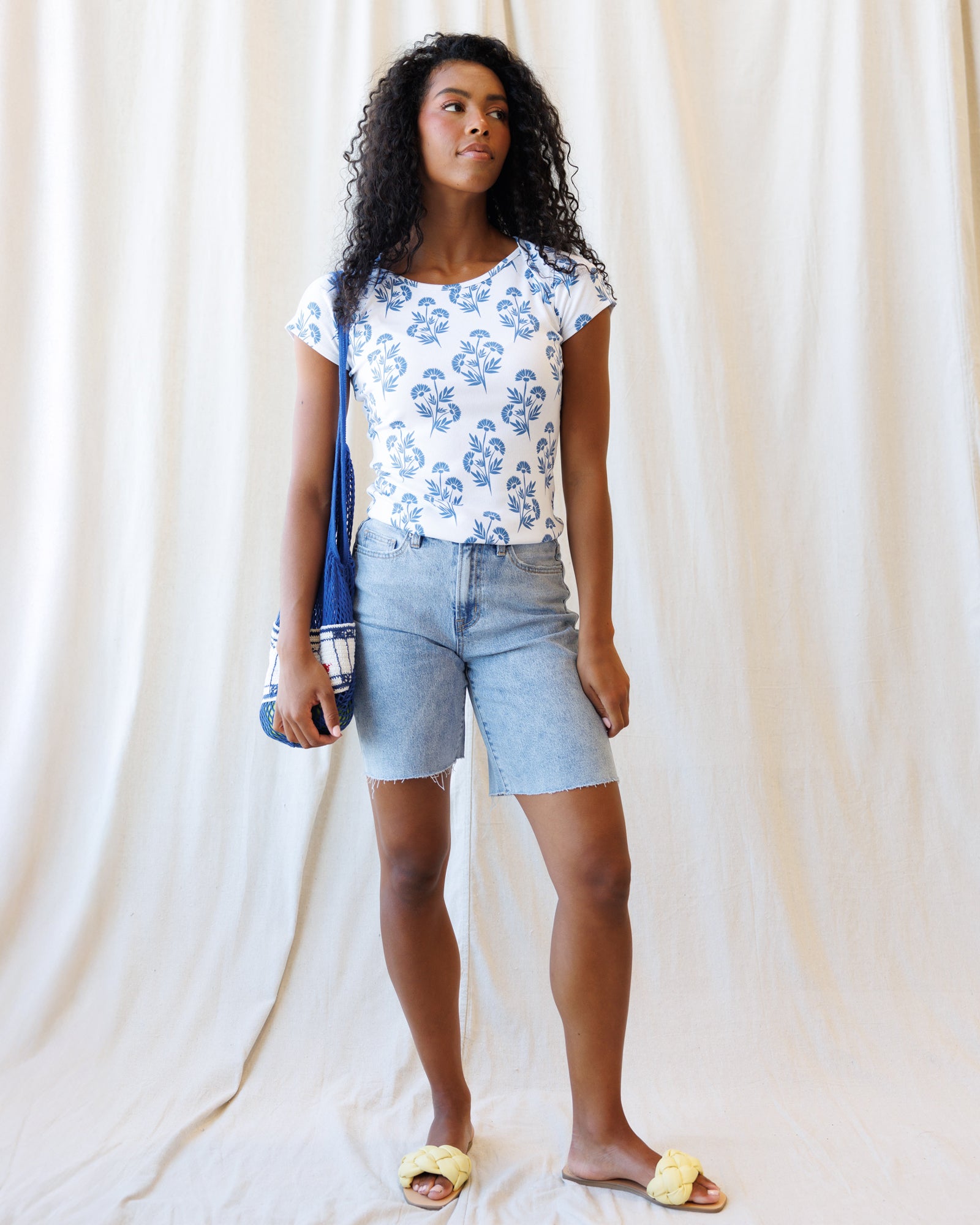 Woman wearing a floral top and denim shorts holding a blue bag against a white curtain background