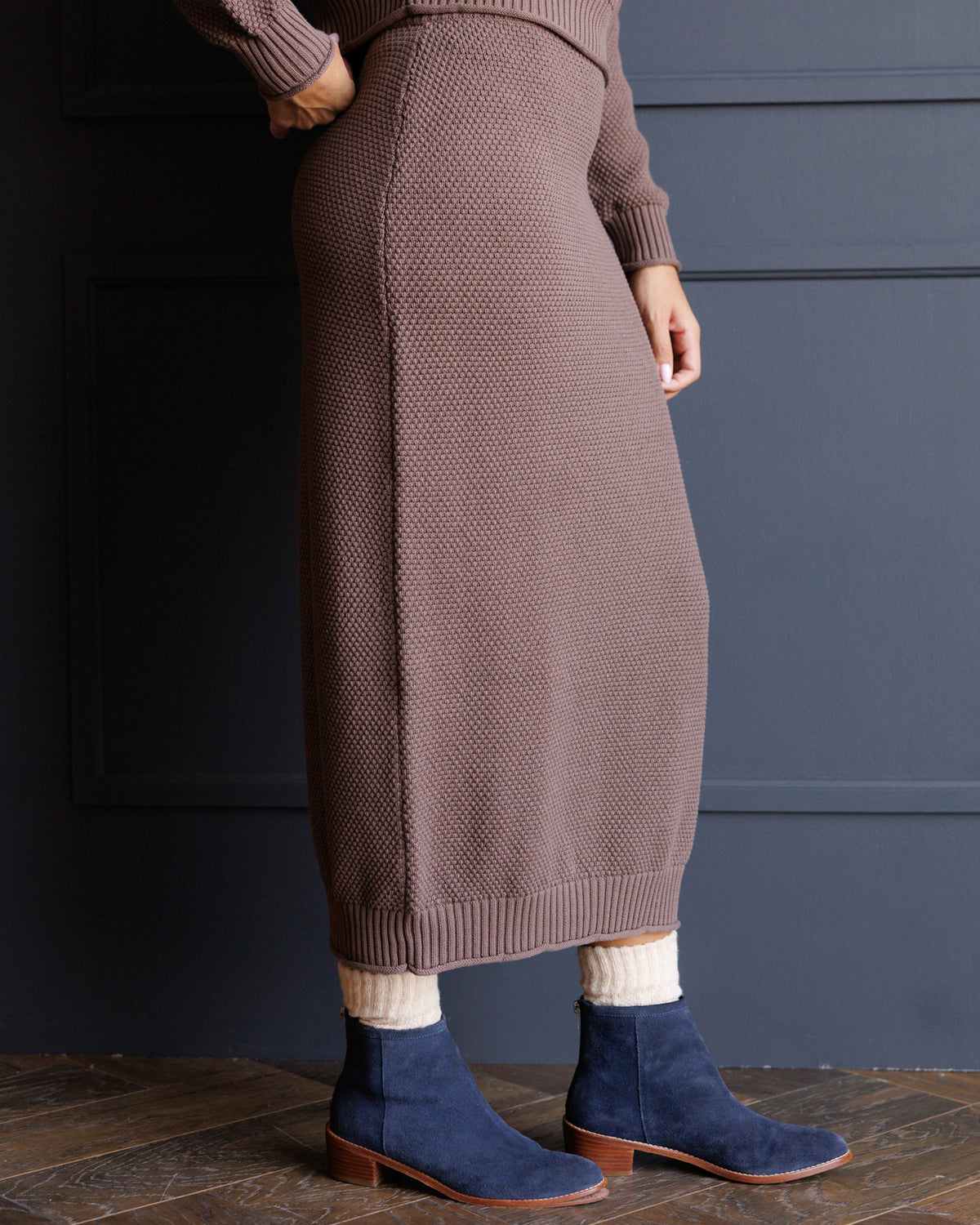 A person wearing a ribbed knit midi skirt in a muted taupe-brown color, paired with navy blue suede ankle boots and cream socks, standing against a dark blue paneled wall.
