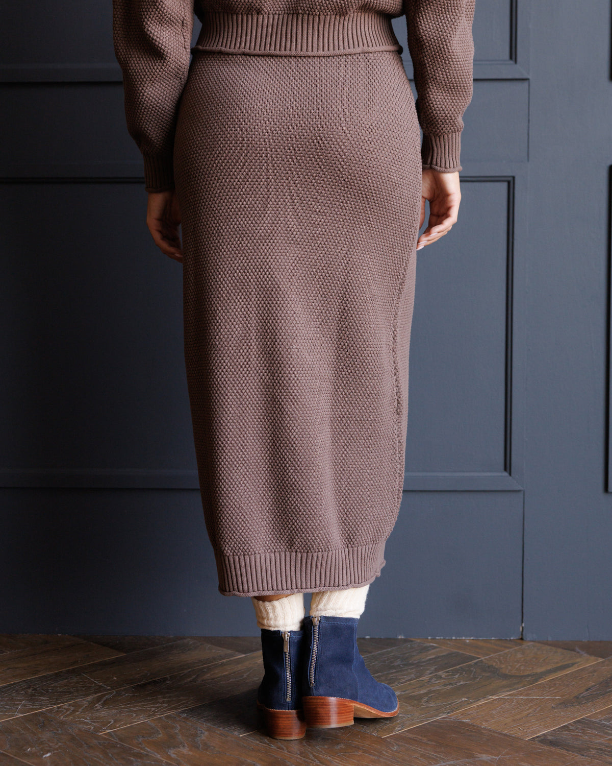 A person shown from the back wearing a ribbed knit midi skirt in taupe-brown, paired with a matching sweater, cream leg warmers, and navy ankle boots, standing against a dark blue paneled wall.