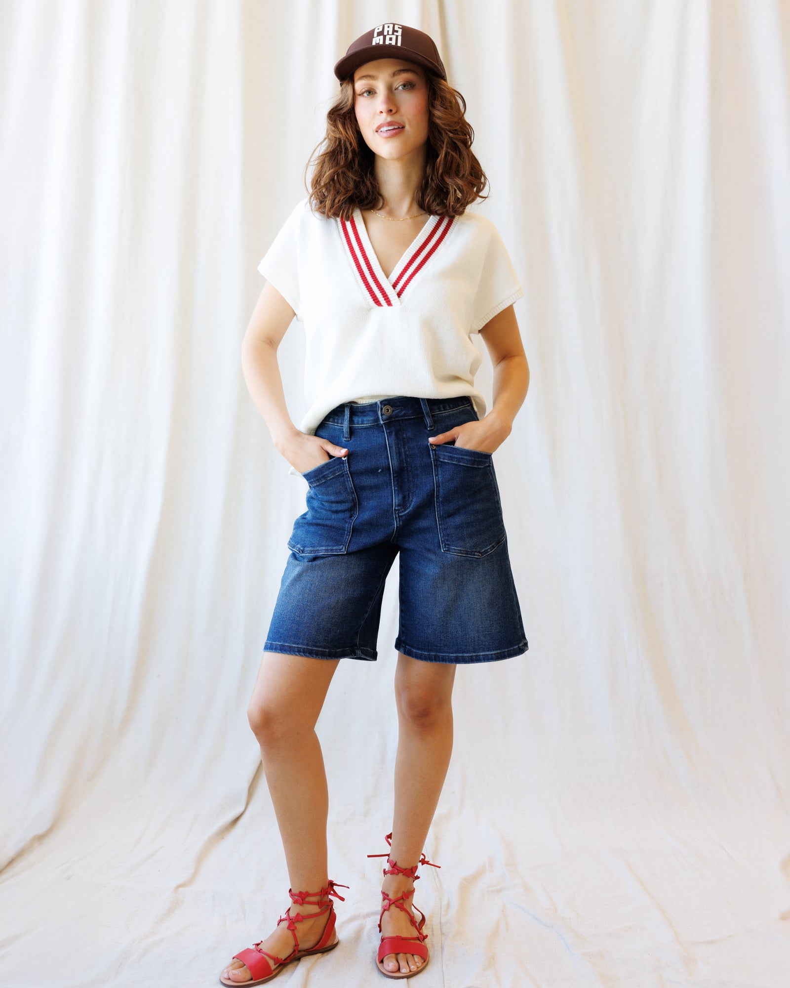 Woman wearing a white V-neck shirt with red accents, blue denim shorts, and pink sandals against a white curtain background.