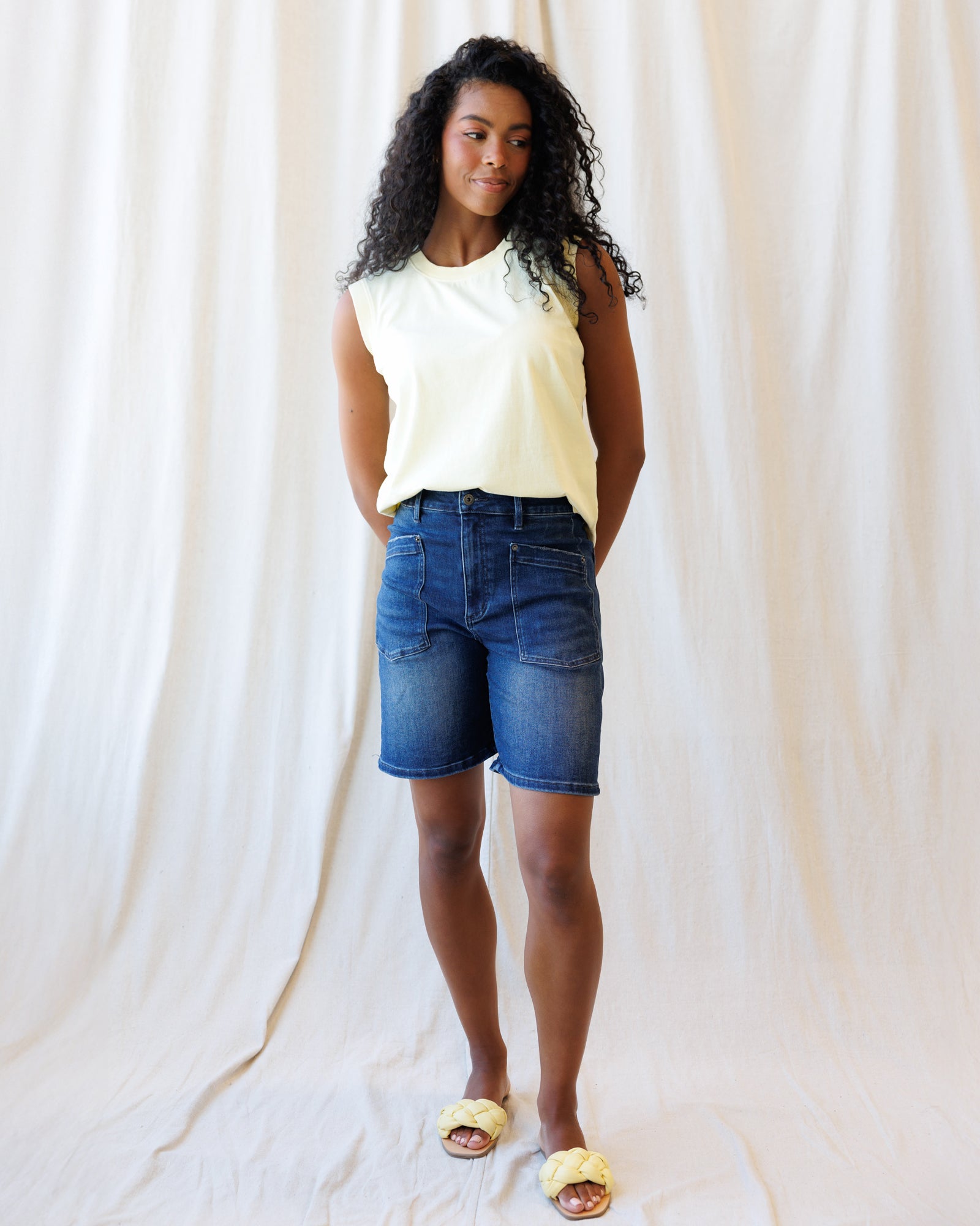 Woman wearing a white sleeveless top and blue denim shorts against a plain background