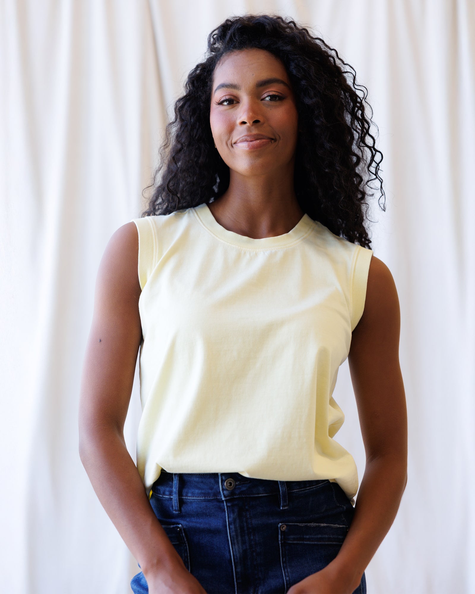 Woman wearing a light yellow sleeveless top and blue jeans against a white curtain background
