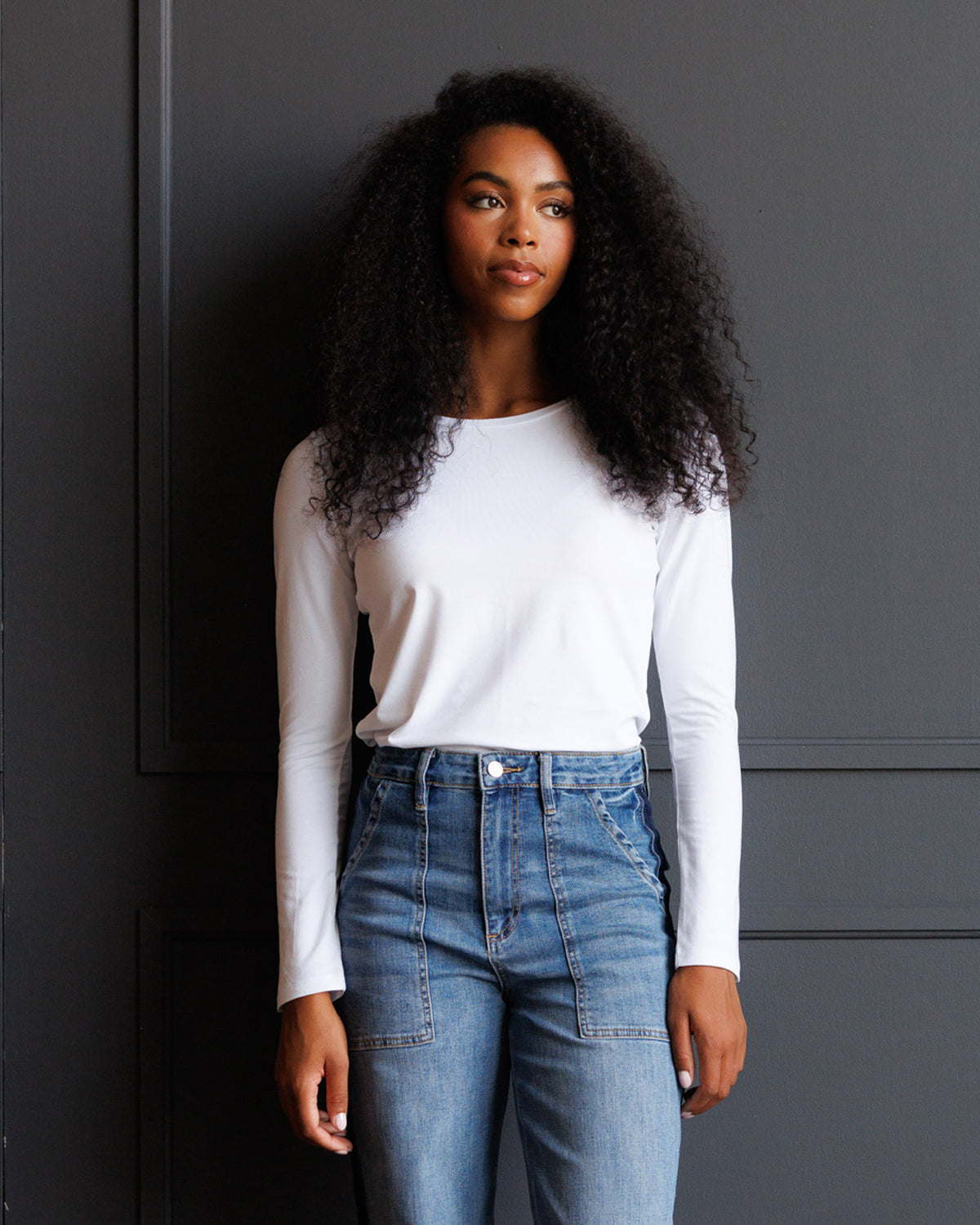 A woman with long, curly black hair stands against a dark gray paneled wall, wearing a fitted white long-sleeve crewneck top and high-waisted blue jeans with unique patch pockets.