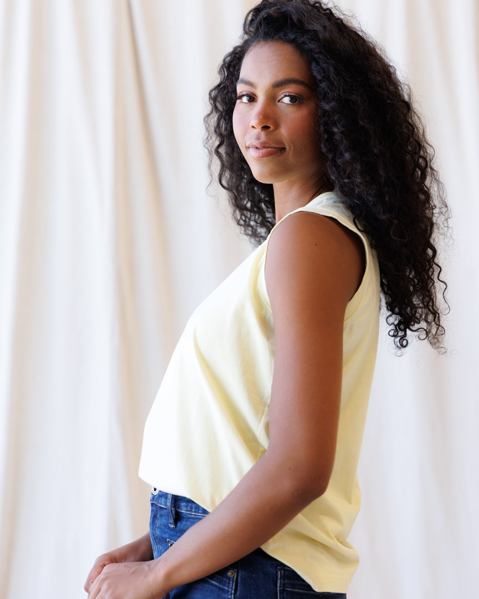 Woman wearing a yellow sleeveless top and blue jeans against a white curtain background