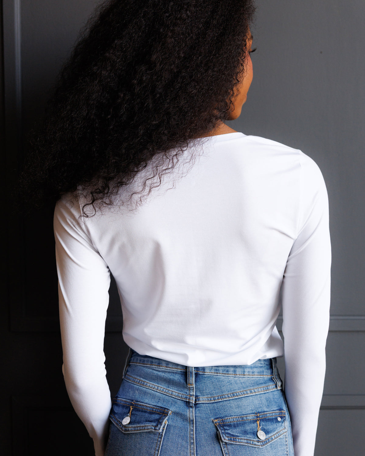 A woman with long, curly black hair is shown from behind wearing a fitted white long-sleeve t-shirt and blue jeans against a dark gray paneled wall.