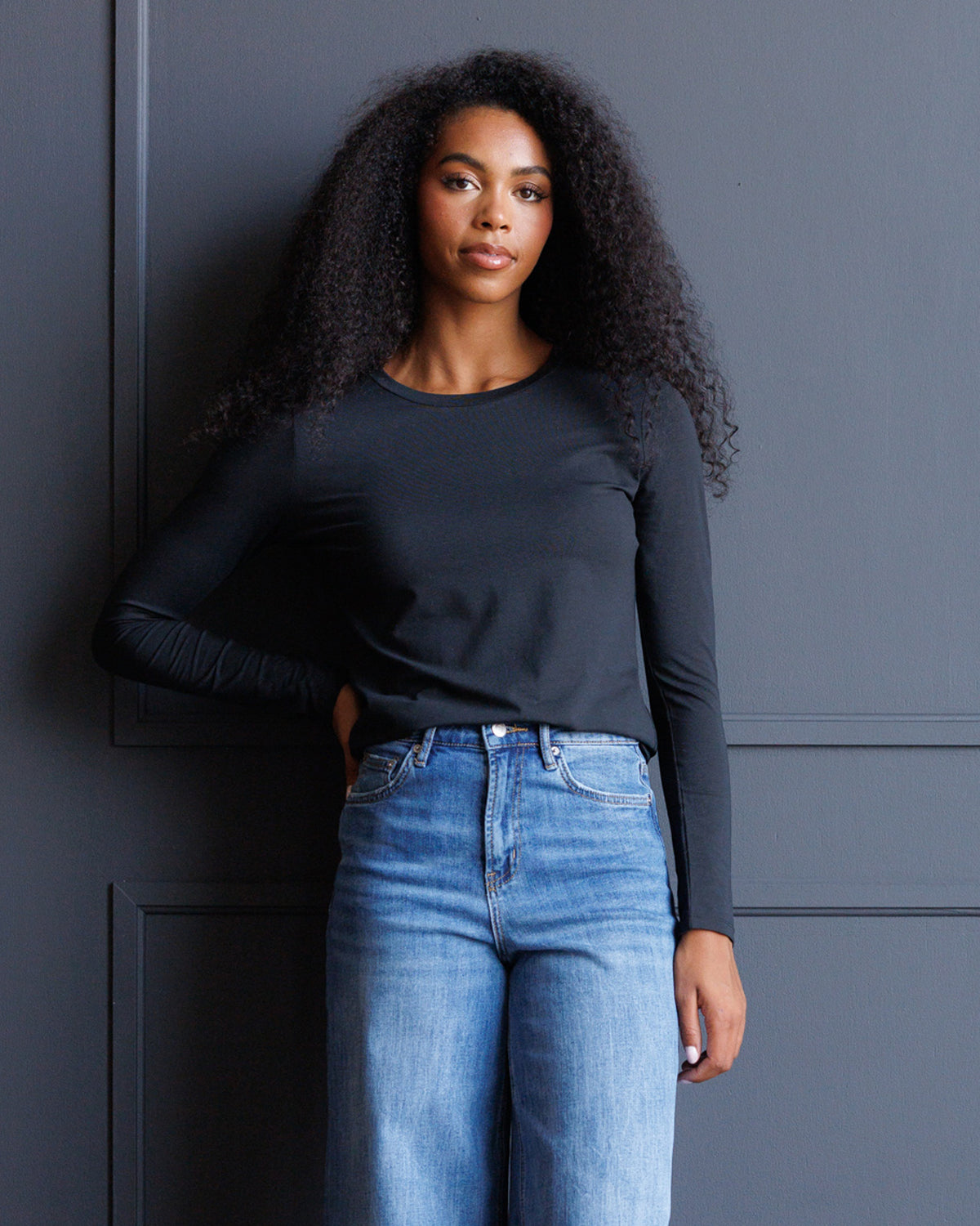 A woman with long, curly black hair stands against a dark gray paneled wall, wearing a fitted black long-sleeve crewneck top and light blue high-waisted jeans.