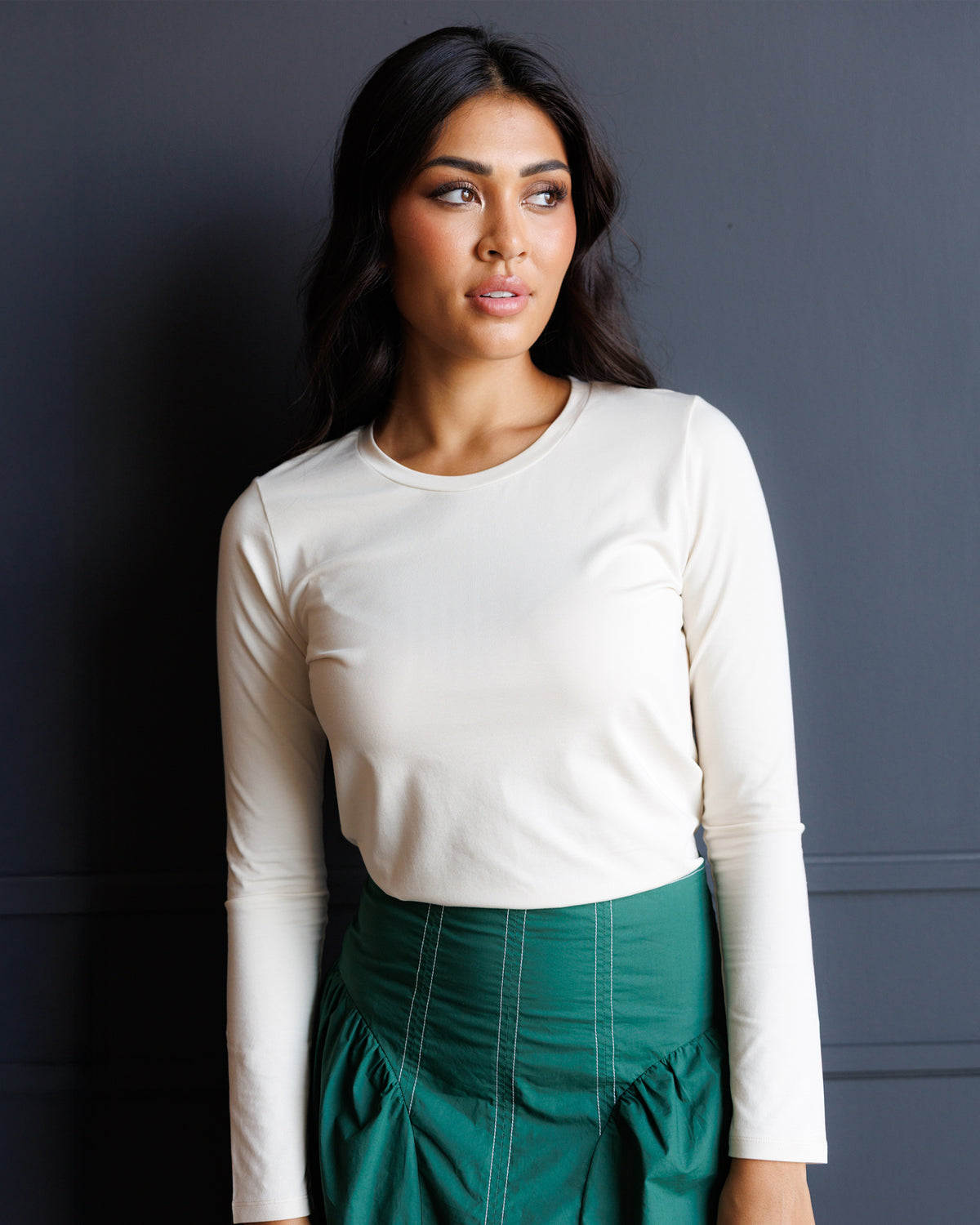 A woman wearing a fitted off-white long-sleeve crewneck top and a dark green high-waisted skirt, standing against a dark gray paneled wall.