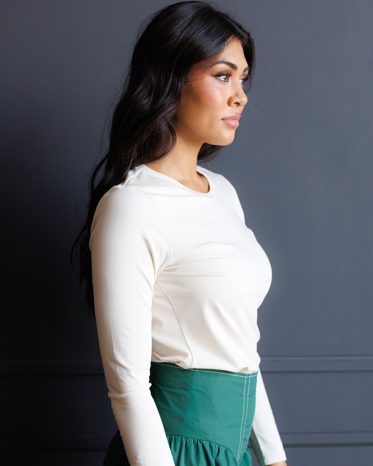 A woman in a side profile wearing a fitted white long-sleeve top and a green skirt, standing against a dark gray wall.