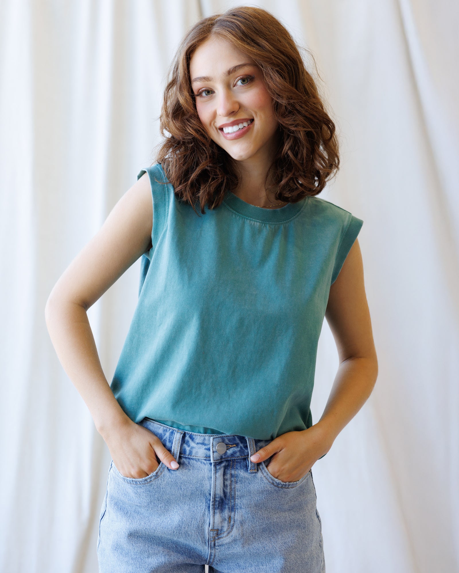 Woman wearing a teal sleeveless top and blue jeans against a white background