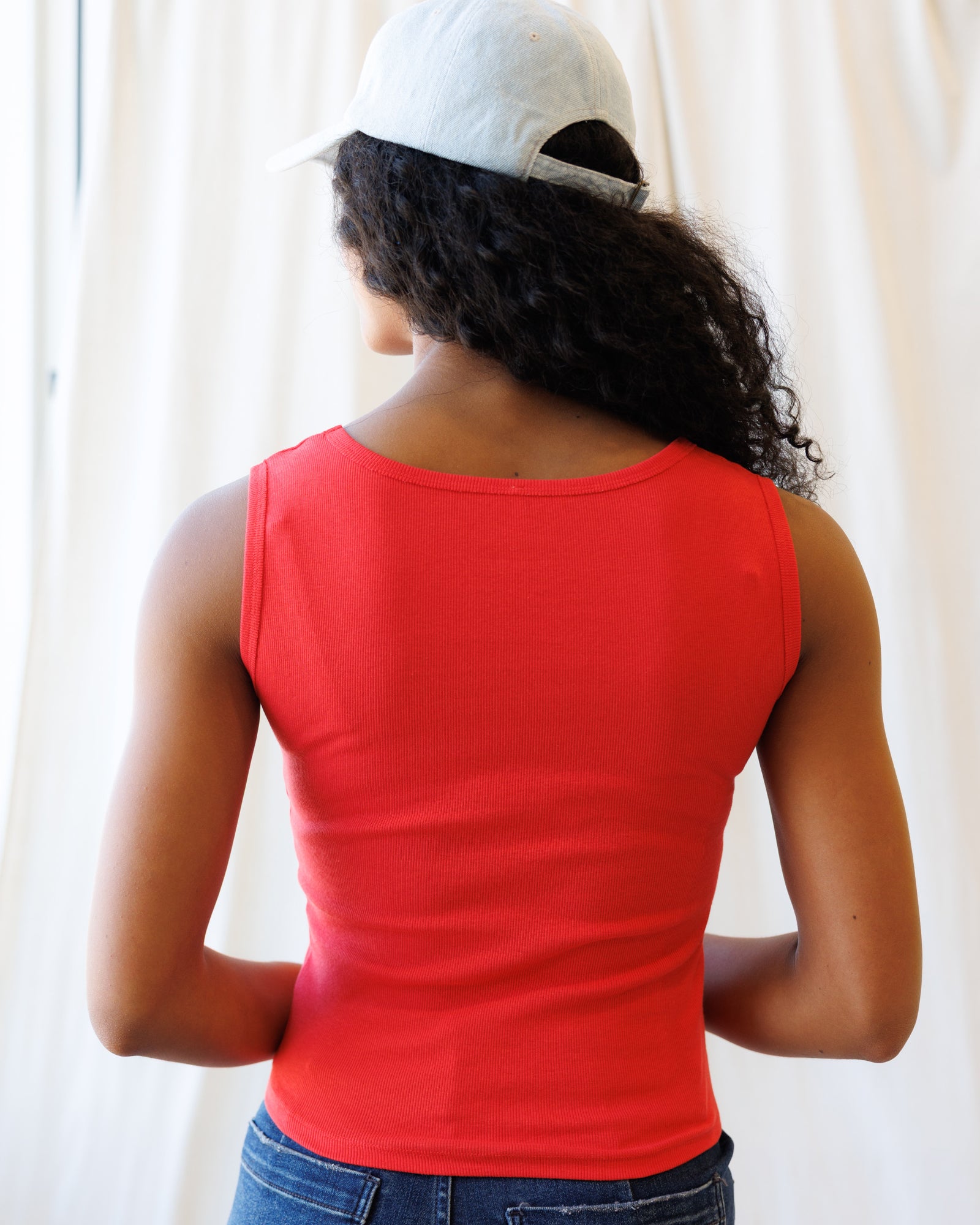 Person wearing a red tank top and white cap from behind, standing against a plain background
