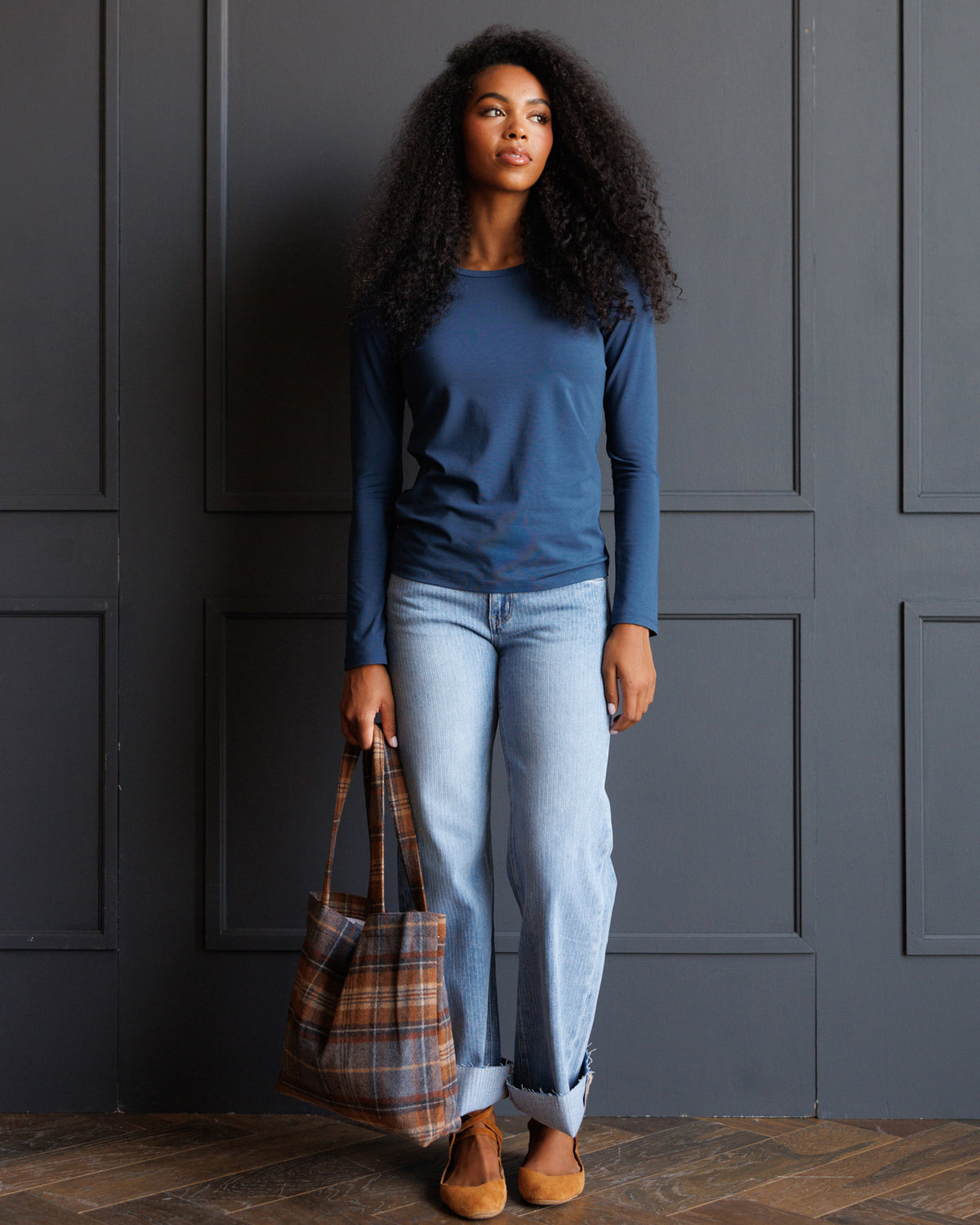 A woman with long curly black hair stands against a dark gray paneled wall, wearing a blue long-sleeve top, light-wash blue jeans, and tan loafers, while holding a brown and blue plaid tote bag.