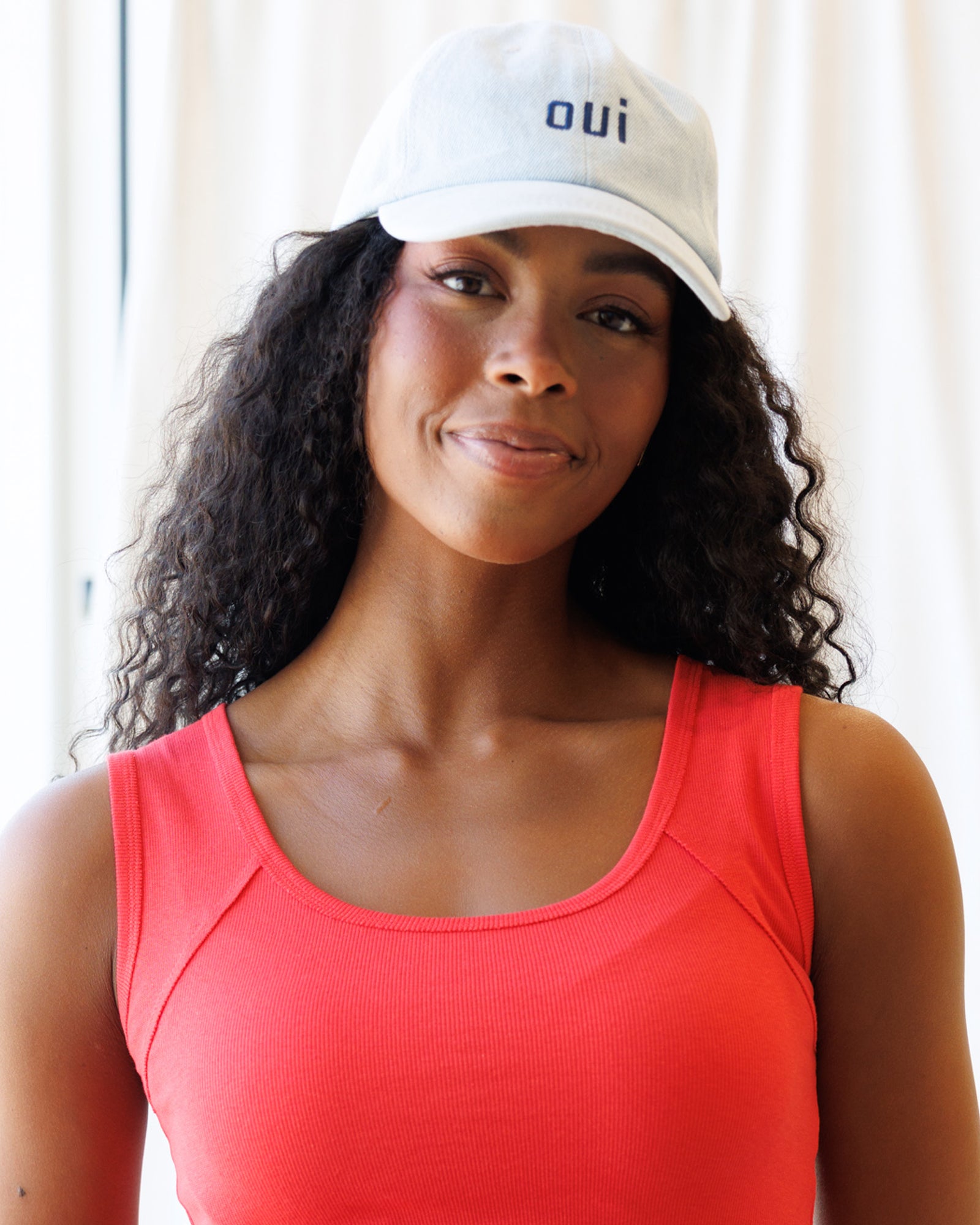 Image of a woman wearing a light wash denim hat with "OUI" embroidered in navy thread and wearing a red tank top