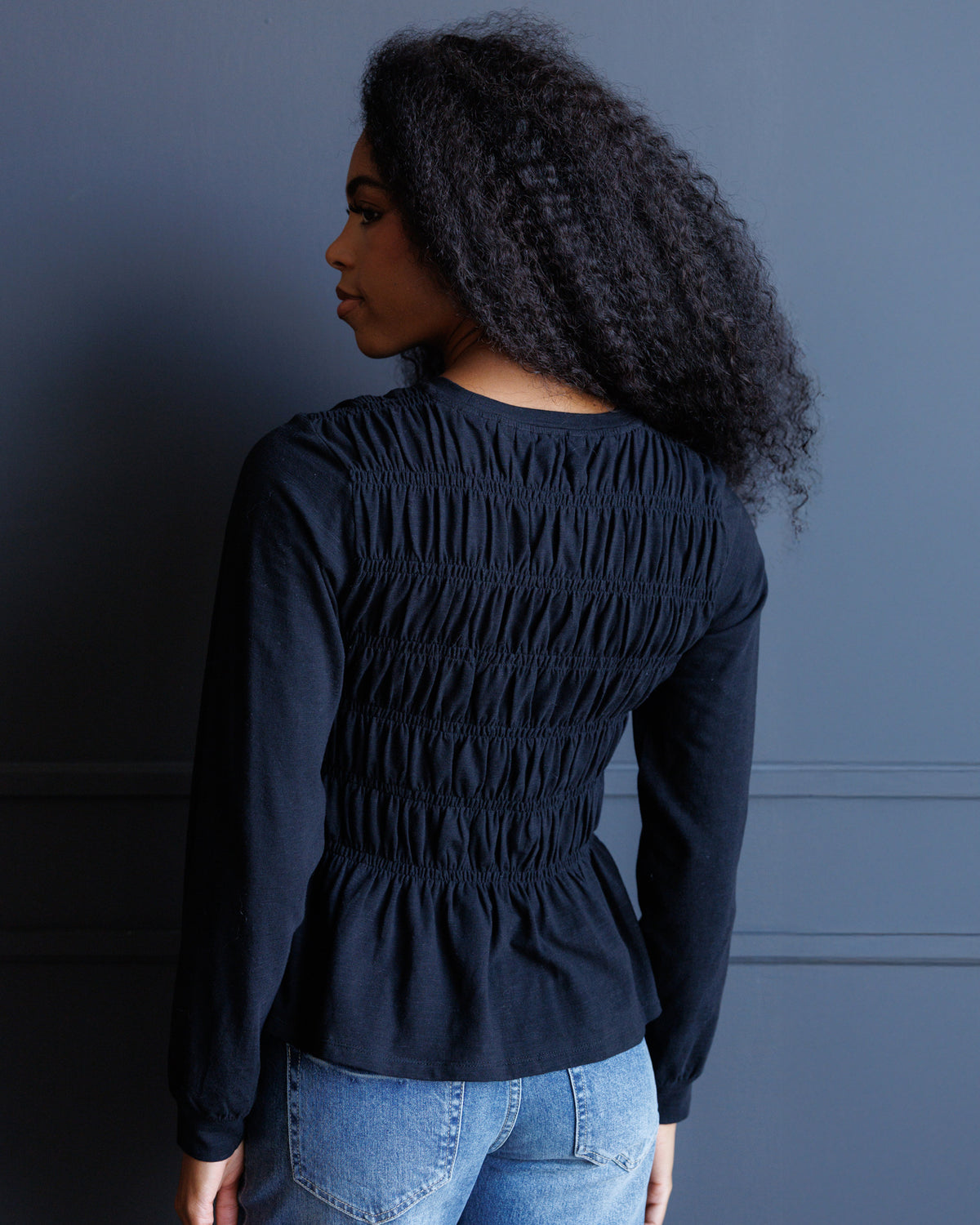 A woman with long, curly black hair is shown from the back wearing a dark navy long-sleeve top with a smocked back and light-wash blue jeans, standing against a dark blue paneled wall.