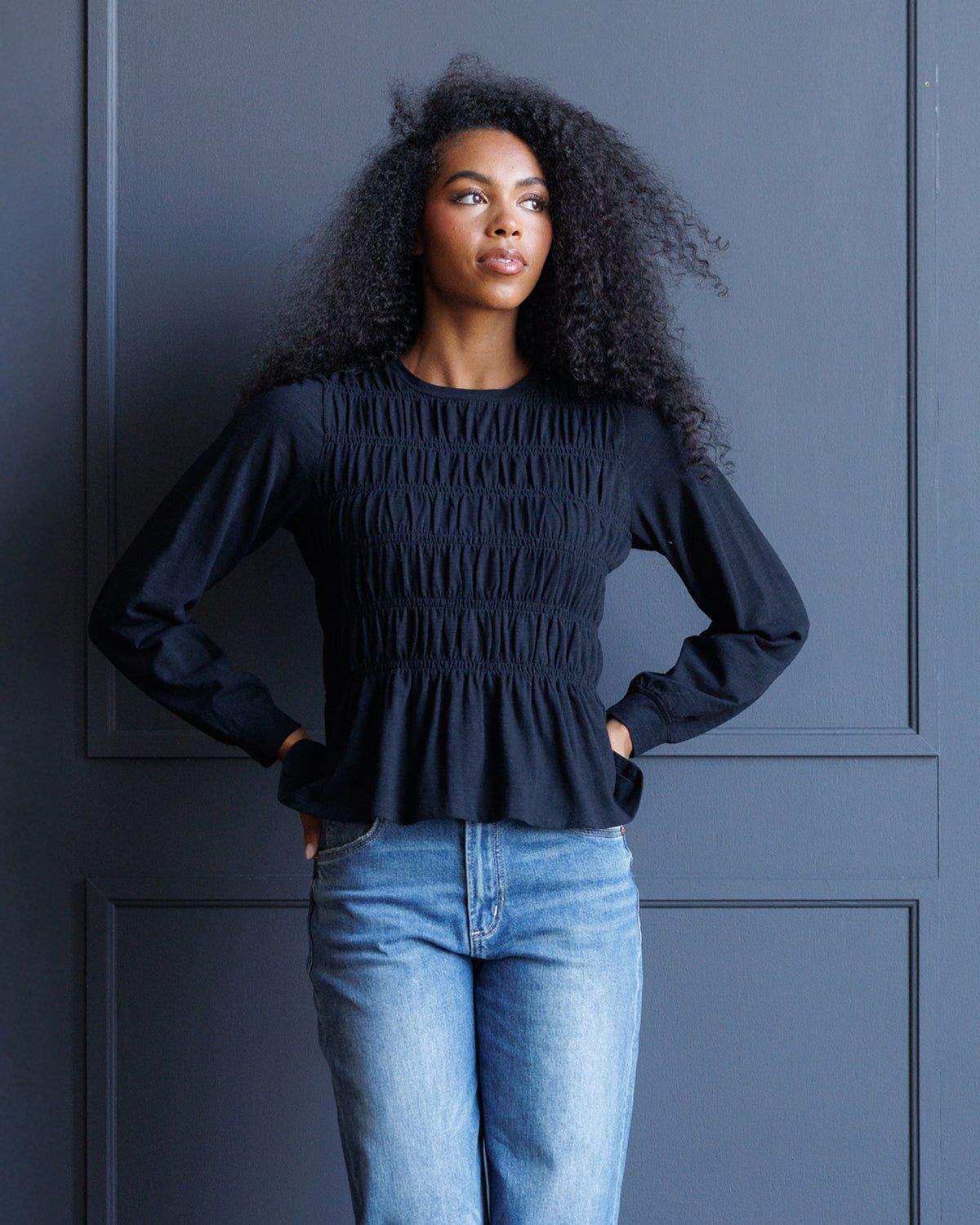 A woman with curly black hair wearing a dark navy long-sleeve smocked blouse and light blue jeans, standing against a dark blue paneled wall.