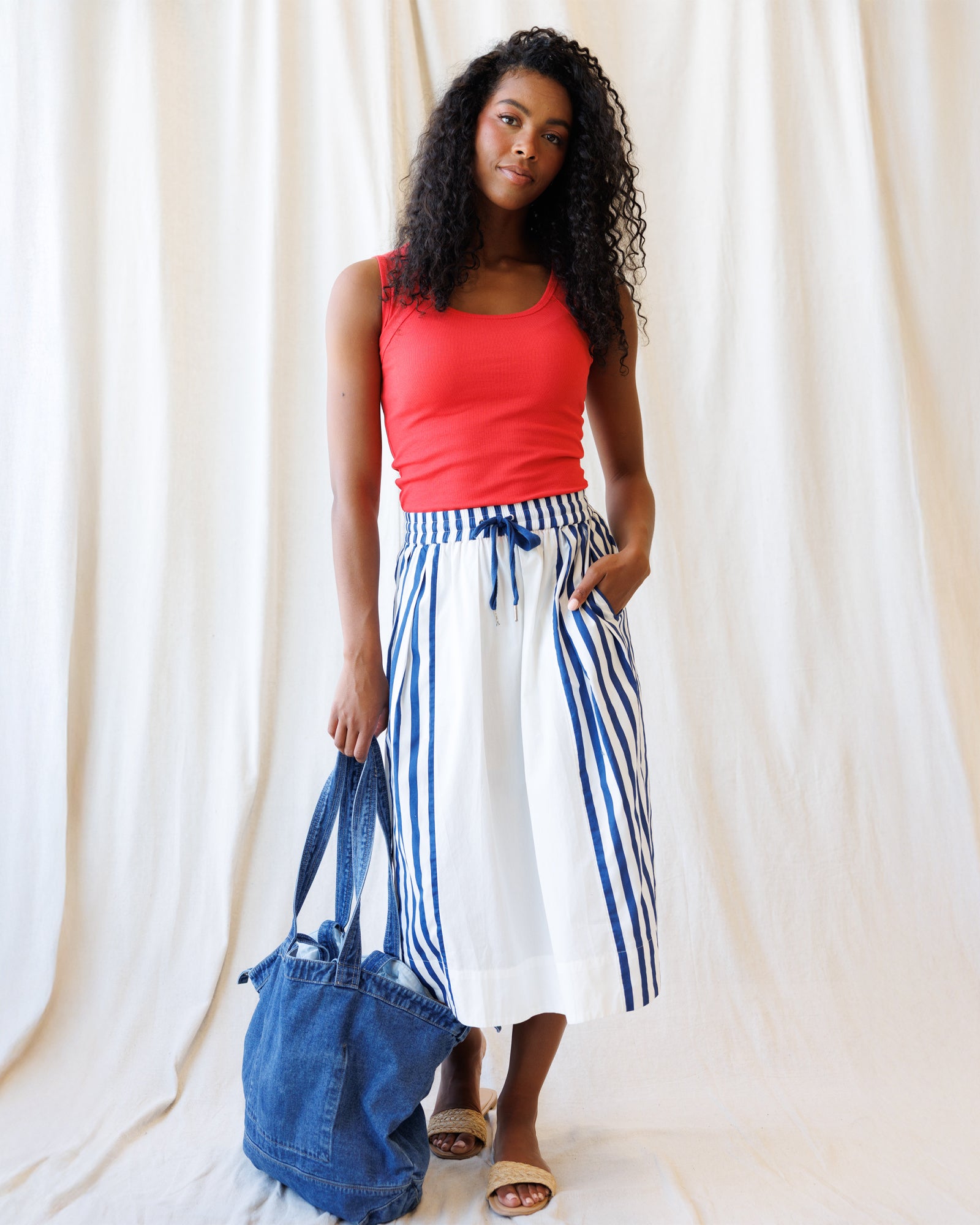 Woman wearing a red top and blue and white striped skirt holding a denim bag against a white curtain background