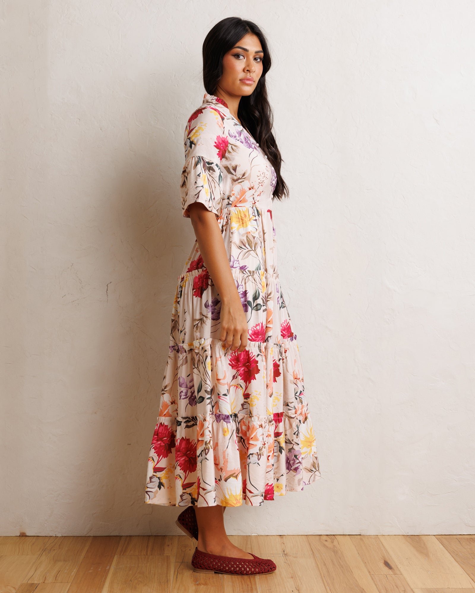 A woman wearing a tiered midi dress with a colorful floral print, standing in profile against a textured off-white wall on a light wood floor.