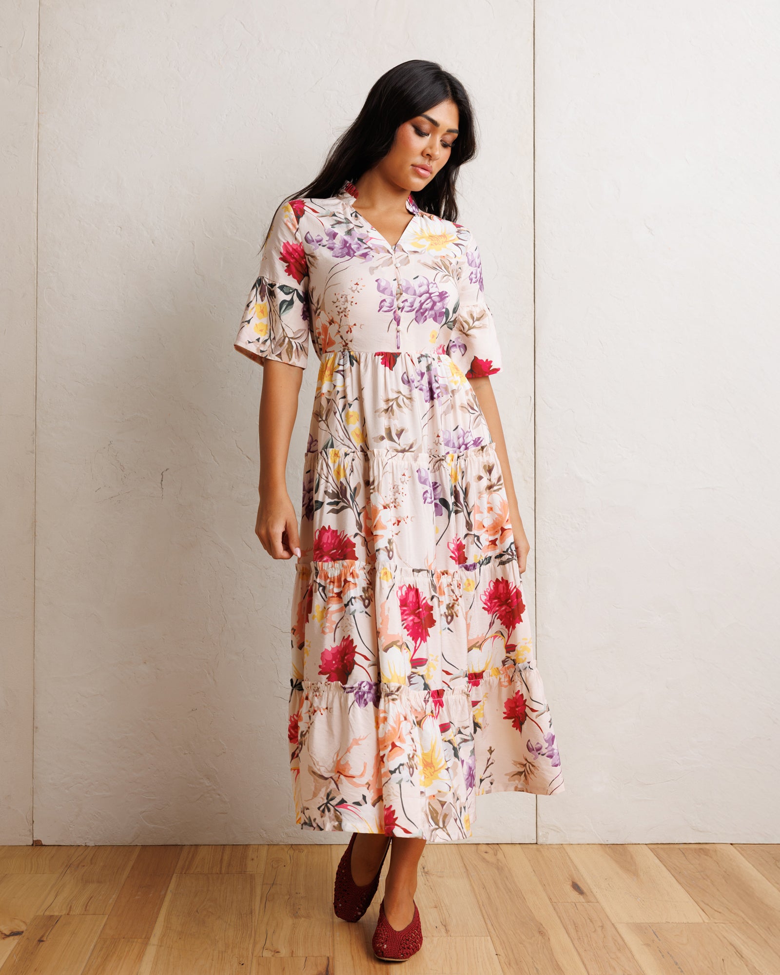 A woman modeling a short-sleeve midi dress with a colorful floral print on a cream background, standing in front of a light-colored wall and wooden floor.