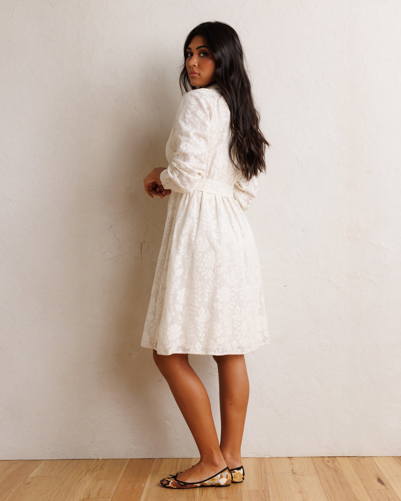 A woman wearing a white textured lace dress with long puff sleeves and a tie waist, standing against a textured off-white wall on a light wood floor.
