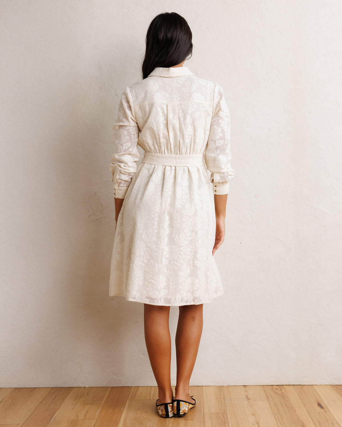 A woman viewed from behind wearing a cream-colored lace long-sleeve shirt dress with a tie waist, standing on a light wood floor against a textured beige wall.