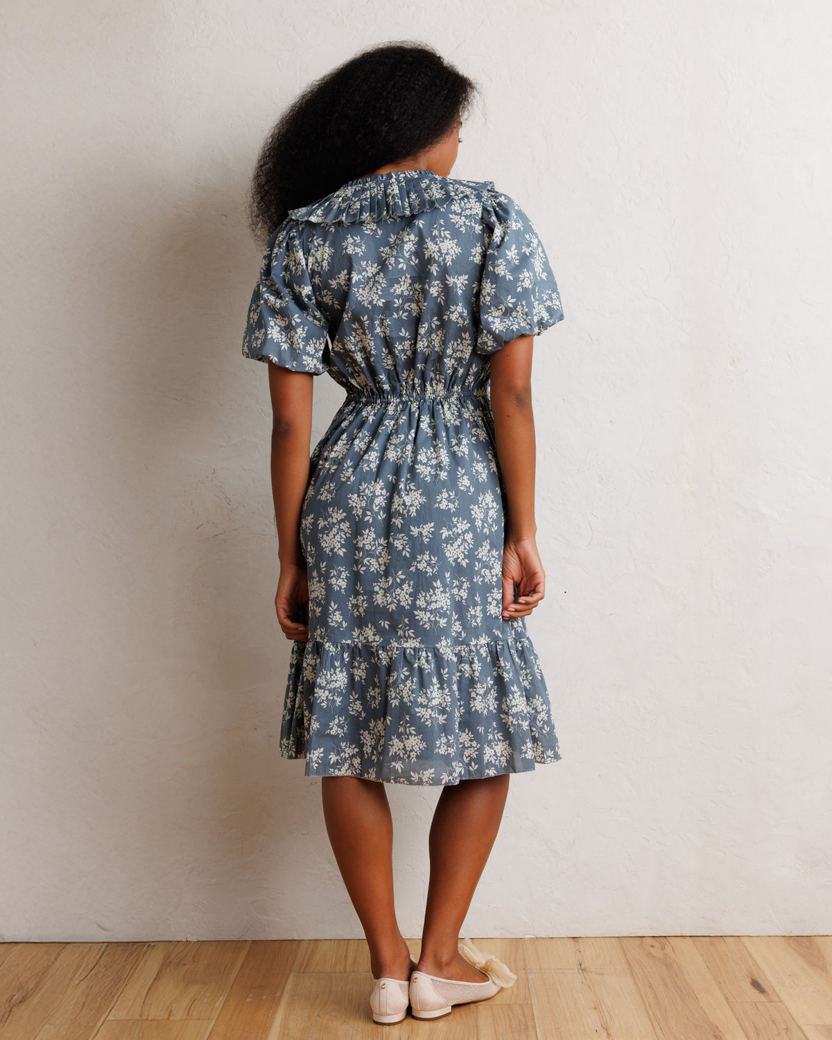 A woman viewed from behind wearing a blue-gray short-sleeve midi dress with a white floral print, gathered at the waist, standing on a light wood floor against a textured off-white wall.