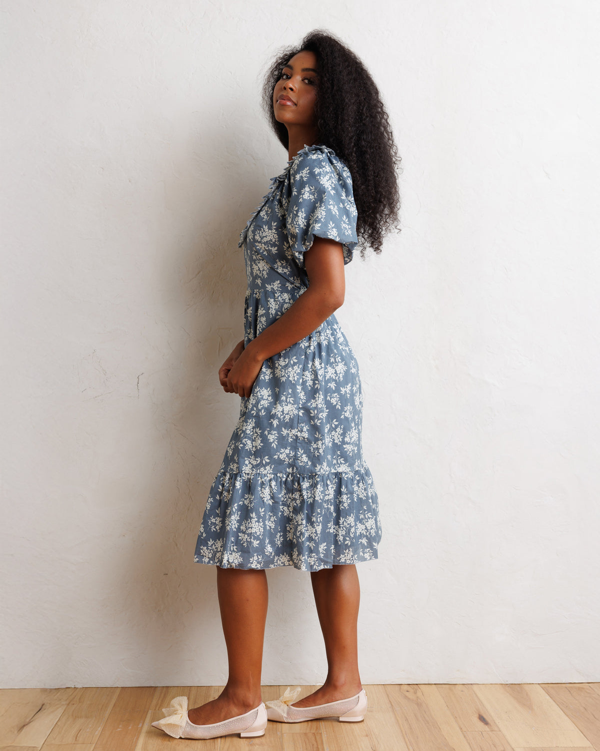 A woman stands in profile wearing a short-sleeve blue floral midi dress with a ruffled hem, paired with pale pink flats, against a plain off-white wall and light wood floor.