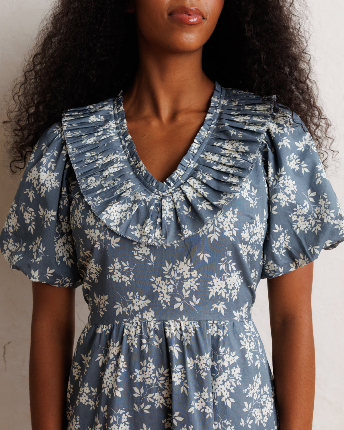 A woman wearing a dusty blue and white floral print dress with a ruffled Peter Pan collar and short puff sleeves, shown from the chest up against a neutral light background.