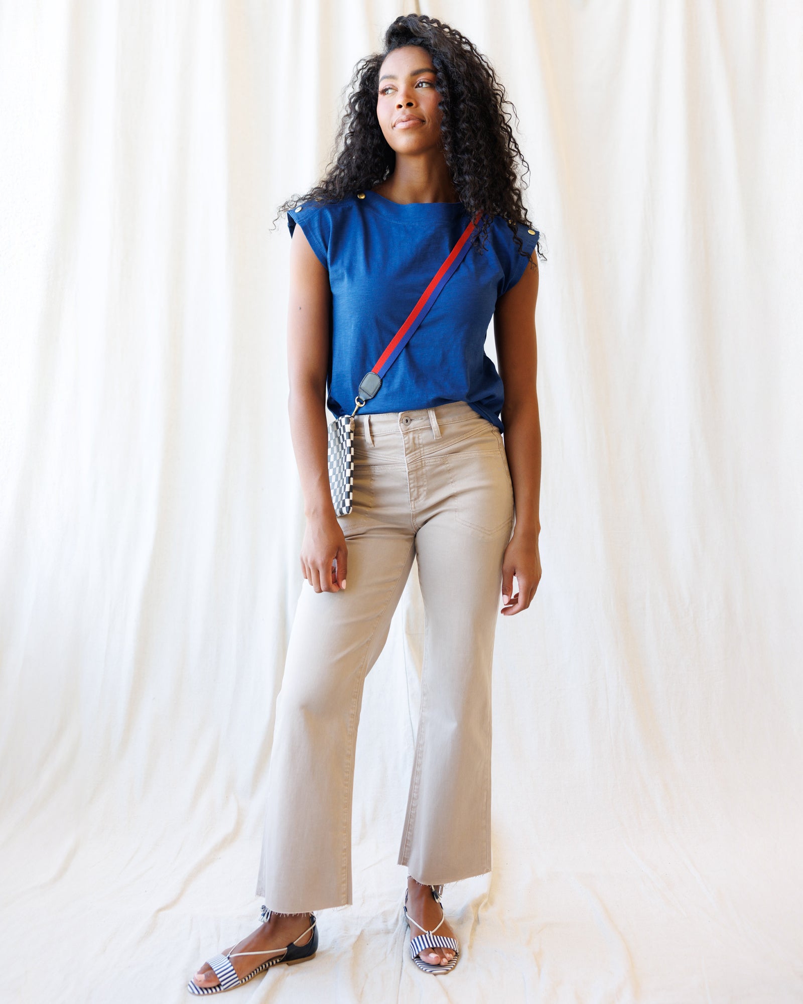 Woman wearing a blue top and beige pants with a red strap over her shoulder against a white background