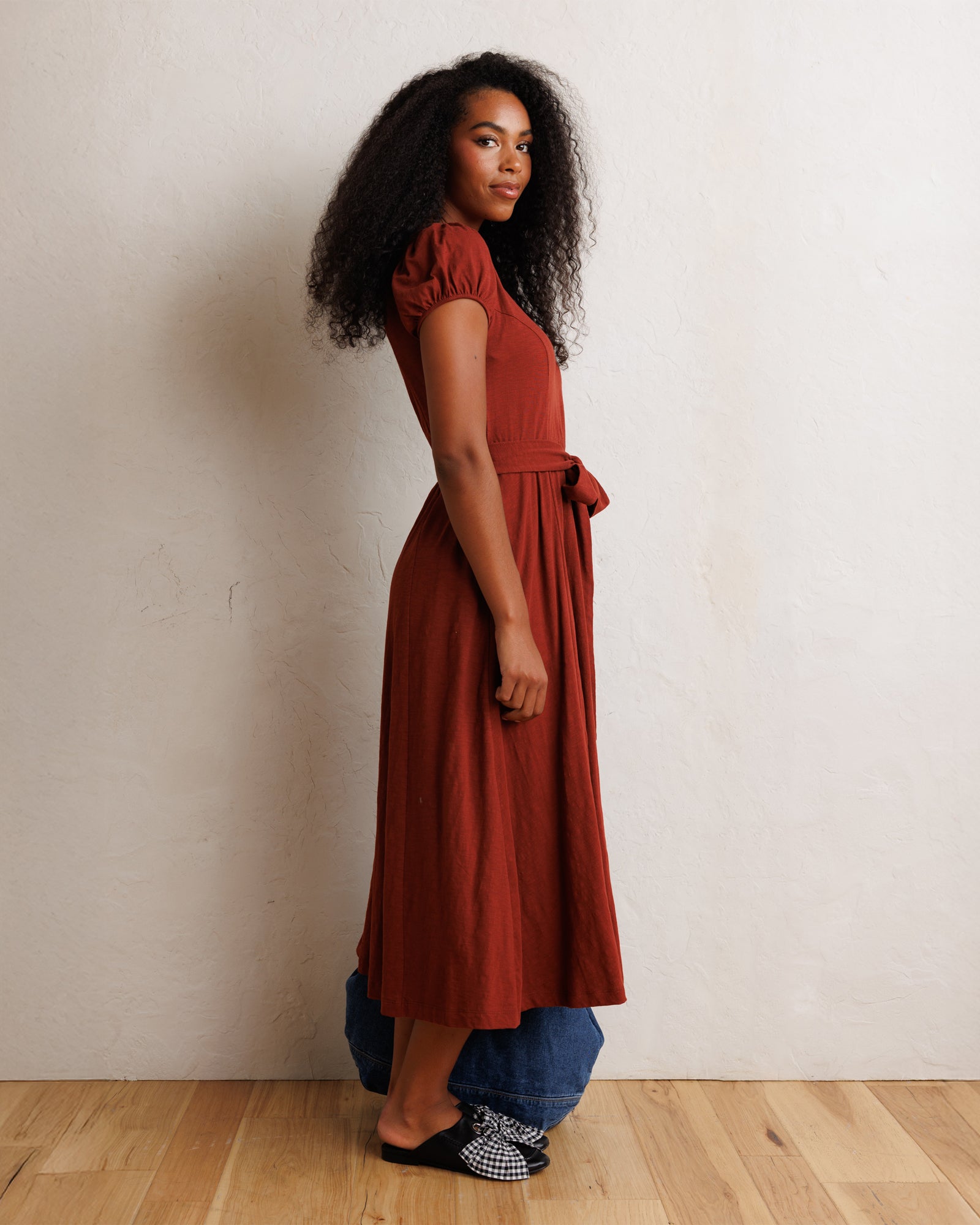 A woman stands in profile wearing a rust-colored midi dress with puff sleeves and a matching belt, layered over blue jeans and black-and-white patterned slip-on shoes, against a textured off-white wall and light wood floor.